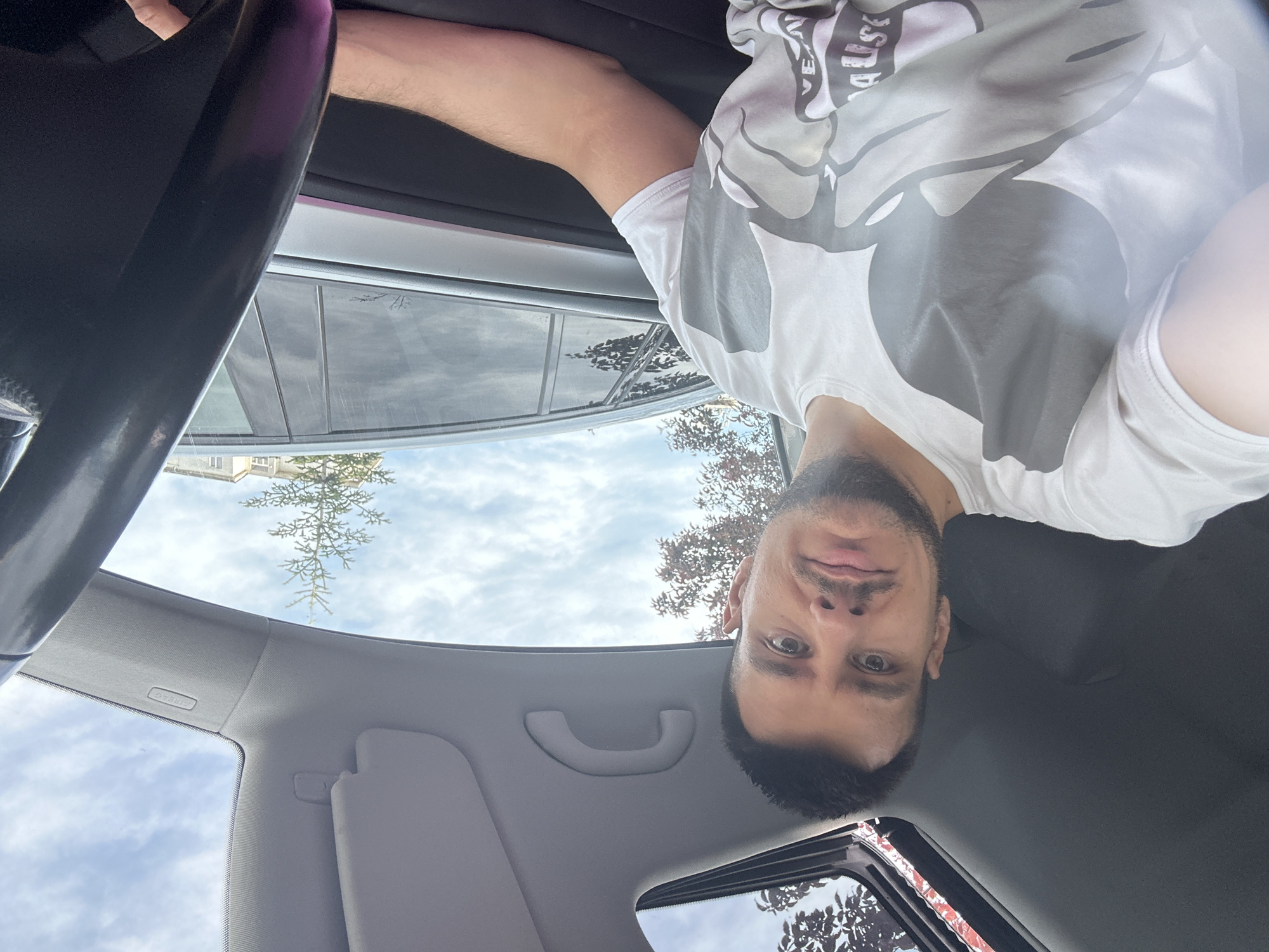 A man is visible inside a vehicle, likely a car, from an inverted perspective. He has a fair complexion, dark hair, and a beard. The man is wearing a light-colored t-shirt with a large, possibly cartoonish, design on the front. The interior of the car is visible, including parts of the steering wheel, dashboard, sun visor, and the car's ceiling. Through the sunroof, a view of a tree and a cloudy sky is seen. Due to the buildings in the reflection on the window, the picture was taken in a city. The location appears to be in Sofia, Bulgaria.