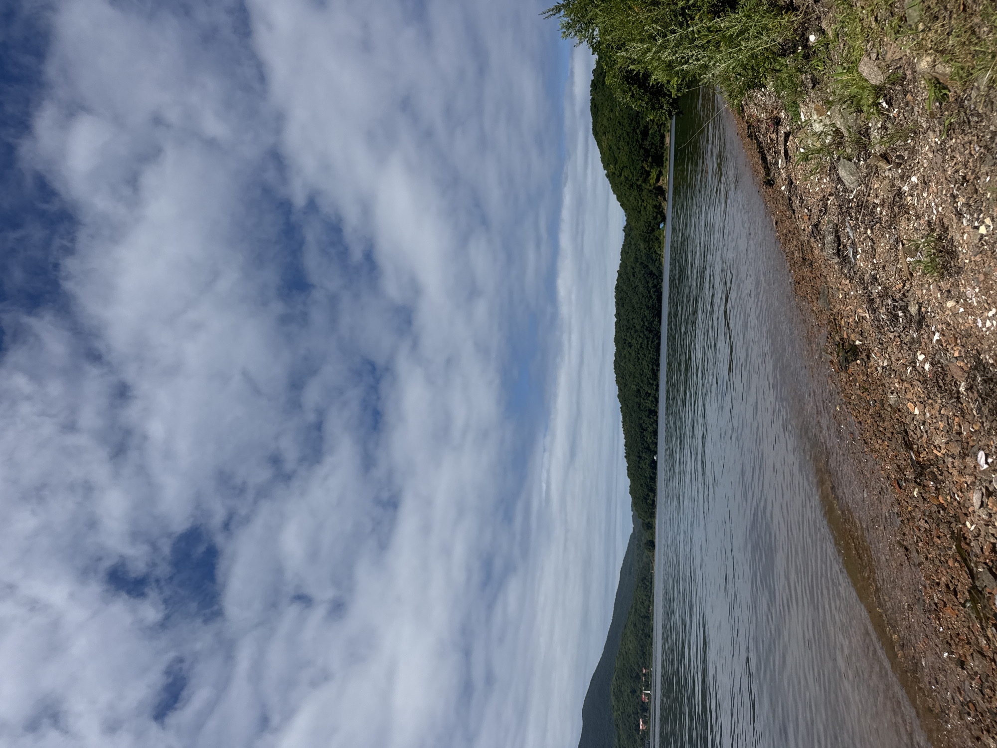 The image depicts a scenic view of a tranquil body of water, likely a lake or bay, under a partly cloudy sky. The foreground features a rocky shoreline with scattered debris and sparse vegetation. The water stretches out towards a distant, tree-covered shoreline with rolling hills or mountains forming the background. The sky is a mix of blue and white clouds, indicating fair weather. There are no people or discernible man-made structures in the immediate foreground or mid-ground, though distant lights or small buildings might be visible on the far shore, suggesting proximity to civilization. The scene is natural and serene, with an emphasis on the landscape and the peaceful atmosphere of a waterside environment. The specific location in Russky, Russia, is not identifiable from the visual information alone.