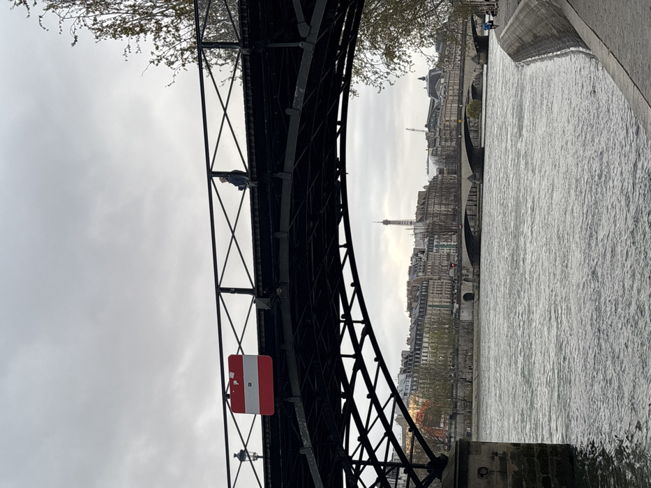 The image presents an overcast daytime scene of the Seine River in Paris, France, viewed from beneath a dark metal arched pedestrian bridge, likely the Pont des Arts, looking generally west-southwest.
The foreground is dominated by the dark, intricate metalwork of the bridge's underside and its pedestrian walkway. A rectangular sign, primarily red with a central white horizontal stripe, is affixed to the bridge's left railing, near a lamppost. On the right side of the bridge railing, a single individual is seated, facing left, dressed in dark attire.
The Seine River occupies the middle ground, displaying a rippled surface. Beyond it, several stone-arched bridges cross the river, with the Pont Neuf prominently identifiable closer to the center. The riverbanks are densely lined with classic multi-story Parisian buildings featuring light-colored facades and grey roofs.
In the distant background, the Eiffel Tower is visible, rising above the urban skyline, slightly to the left of the image's vertical center. On the far right riverbank, a paved walkway runs parallel to the river, where a lone individual is observed jogging towards the right. Trees with sparse, light-colored foliage are present along the riverbanks. The sky is uniformly grey and overcast, indicating a cloudy day.