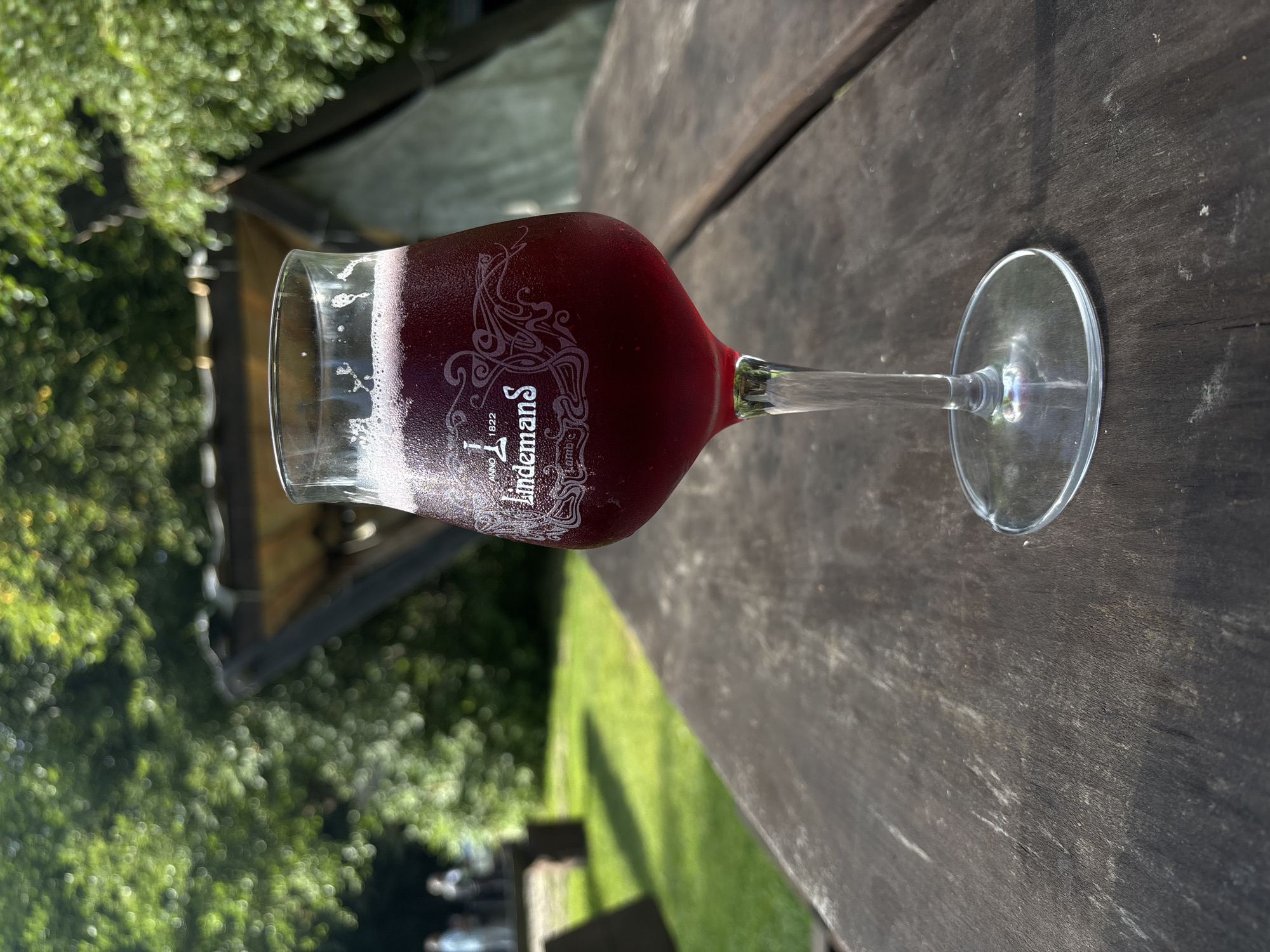 A glass of dark red beverage with a frothy head sits on a rustic wooden table outdoors. The glass is frosted and has the Lindemans logo etched onto it, along with "ANNO 1822" and "Lambic". In the background, green trees and foliage are visible, as well as a blurred structure that appears to be a wooden roof or shelter. A patch of green grass and a picnic table with figures in the distance suggests an outdoor seating area, possibly a park or a restaurant with an outdoor garden. The overall lighting indicates it is daytime, likely a sunny day. The location is not identifiable as Saula, Estonia based on the visual information provided.