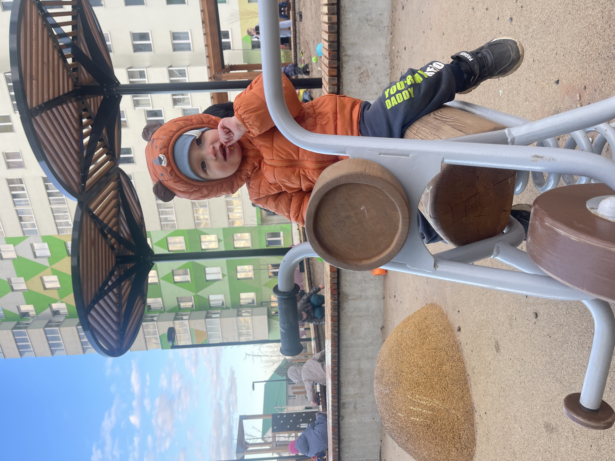 A young child wearing a rust-colored hooded jacket with bear ears, a blue hat, and black pants with "You're My Daddy" written in yellow, is sitting on a spring rider at a playground. The spring rider is made of grey metal with wooden features. The child is facing the camera and has a finger in their mouth. In the background, there's a multi-story building with green, yellow, and white patterned siding. Other playground equipment and people are faintly visible in the background. Based on visual cues such as the architecture and park design, the location is identified as Izhevsk, Russia.