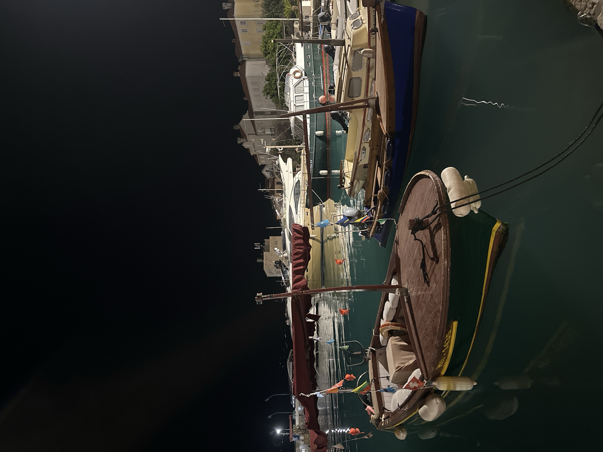 The image, captured at night, displays a serene harbor scene in Budva, Montenegro. The dark night sky dominates the left side, contrasting with the illuminated waterfront and boats on the right. Several vessels are moored in the calm, dark water, which reflects the artificial lights from the shore.
In the foreground, a traditional-style wooden boat is prominently featured. It has a brown deck, a green hull with a yellow stripe, and is adorned with colorful small flags (bunting). White cylindrical fenders are visible along its side. A dark red sail or cover is gathered on a mast, while a life preserver can be seen within the boat. The registration "HR-1435" is visible in yellow lettering on its hull.
Behind this boat, other vessels are docked, including a larger white yacht and a boat with a green hull and a white cabin. Buildings with light-colored facades and red roofs line the distant shore, characteristic of a coastal town. Streetlights or dock lights cast their glow, creating wavy reflections on the water. The scene portrays a peaceful, quiet evening in the marina, with boats at rest.