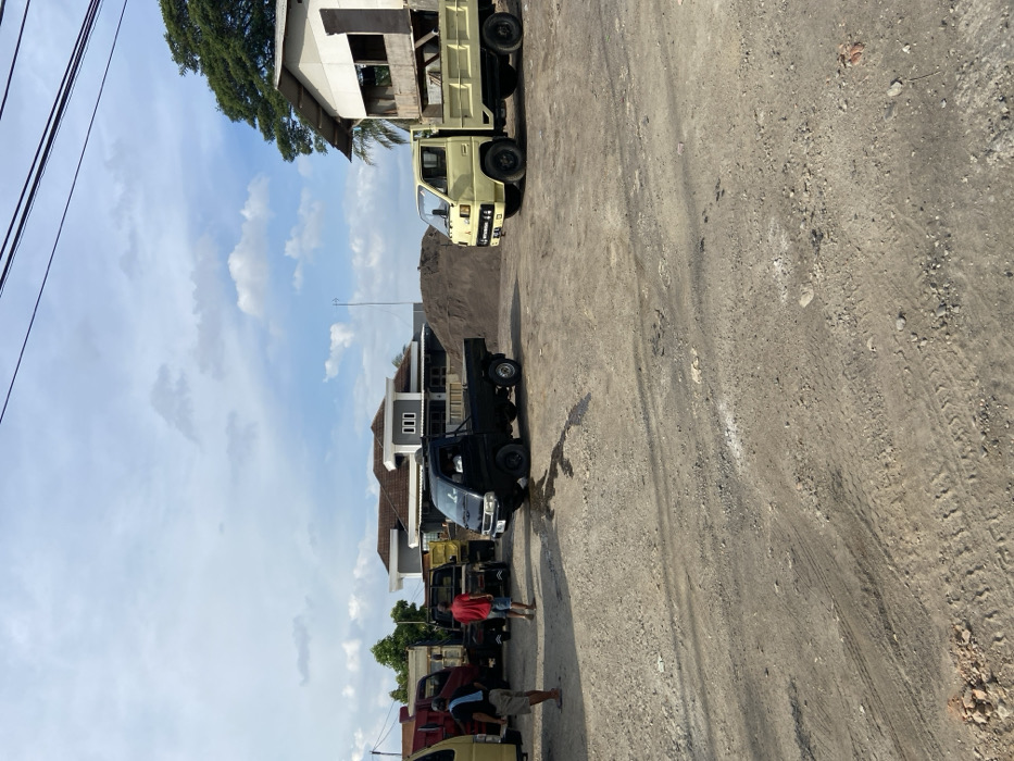 The image shows a dirt road in Jatiluhur, Indonesia, with four trucks parked along the side. Two men are standing to the left of the image. The trucks are facing towards a pile of dirt, likely for construction. The image shows a partly cloudy sky. The trucks are parked along the side of a dirt road. The dirt road is dusty and uneven. The trucks are all old and battered. The men are wearing casual clothing.