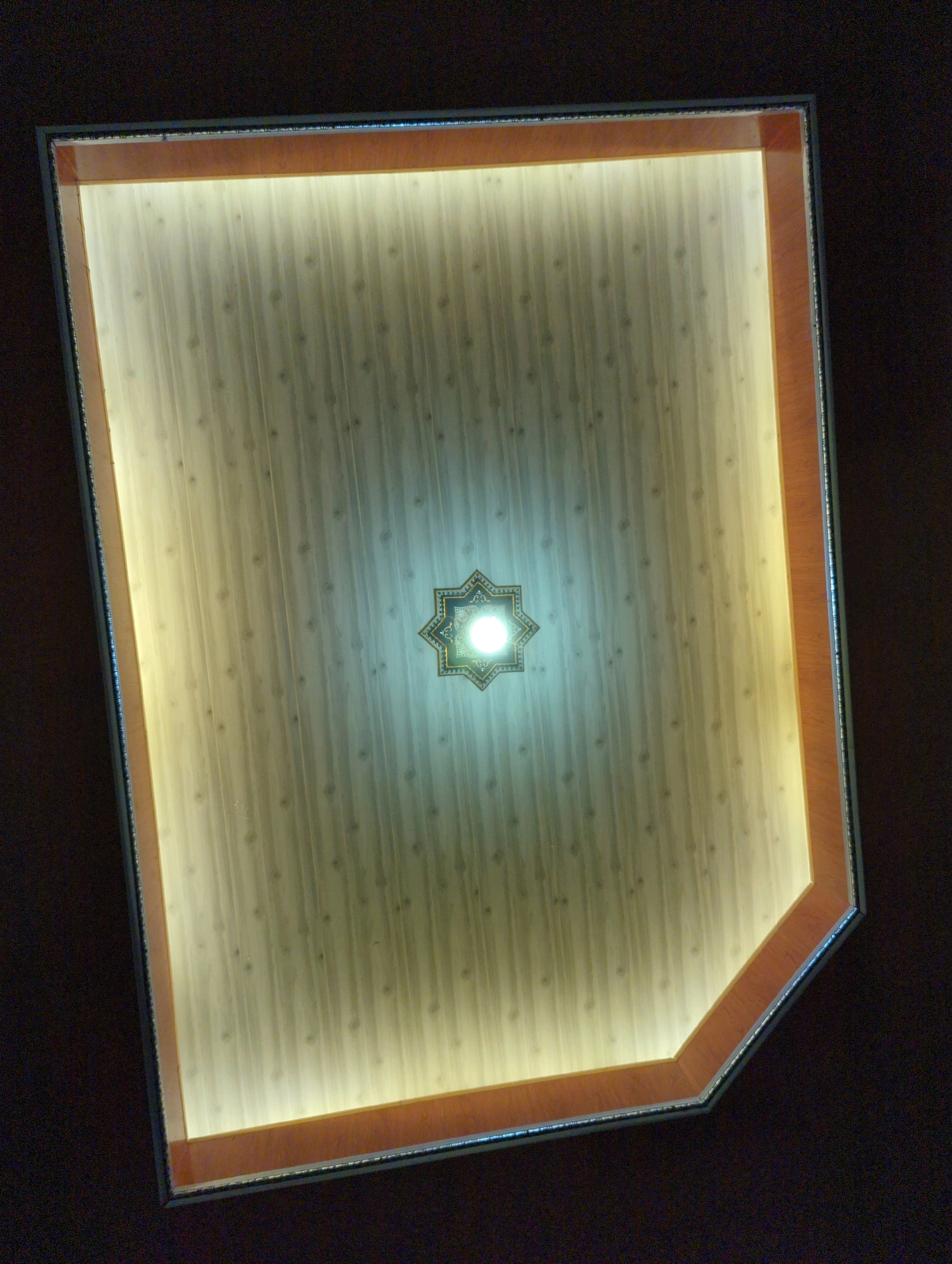A high-angle shot captures a decorative ceiling featuring a central, illuminated star-shaped fixture. The ceiling itself is paneled with a light-colored material that has vertical striations and a subtle pattern of small, circular motifs. A warm, indirect light emanates from behind a decorative border, casting a soft glow on the ceiling panels. The border is composed of an outer metallic trim and an inner wood-toned frame with a beveled edge. The surrounding area is dark, suggesting a dimly lit room. The image's perspective suggests it was taken from below, looking upwards. The image does not provide any identifiable landmarks or context to place it within Kedungdowo, Indonesia.