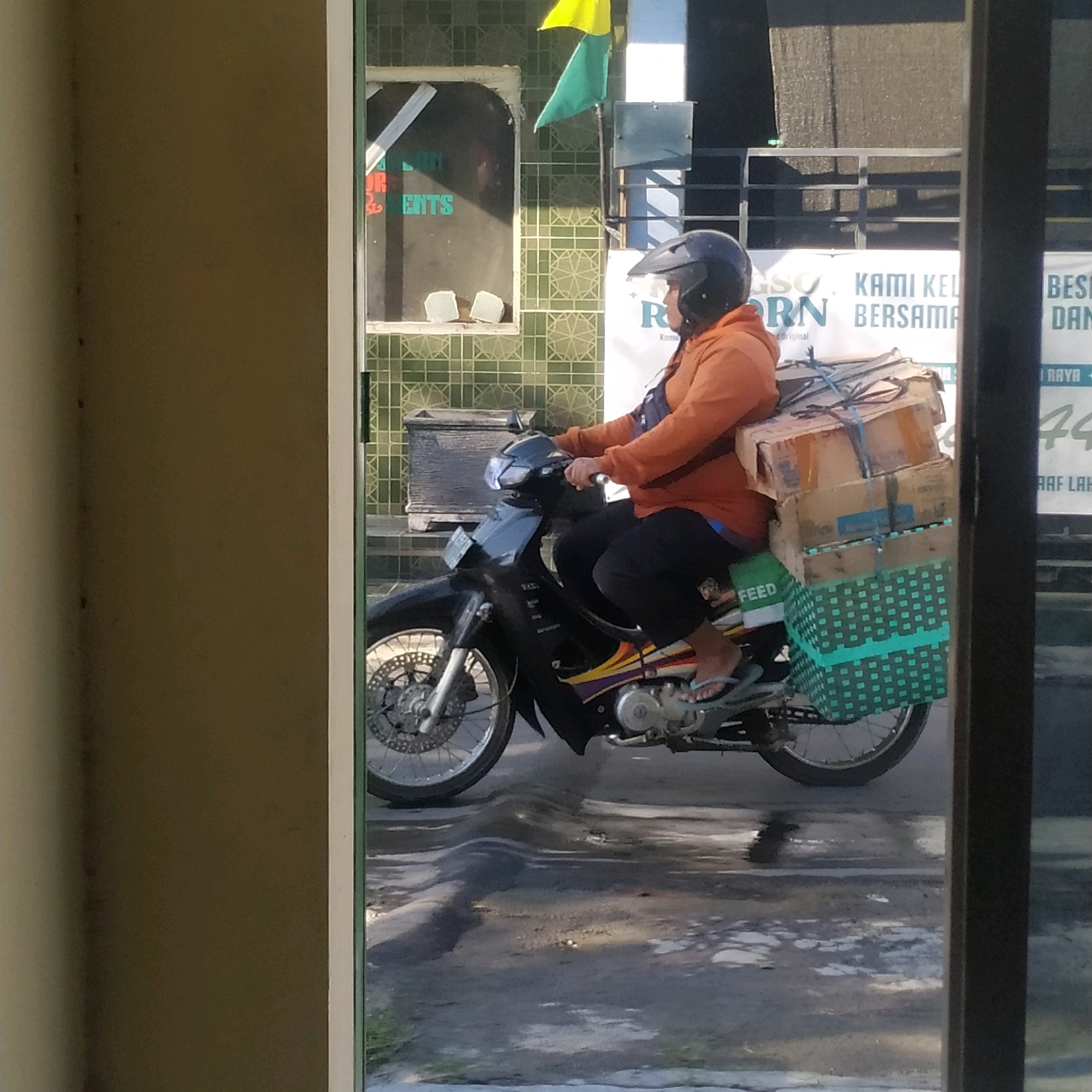 A side profile of a person wearing a black helmet and an orange long-sleeved shirt is riding a black motorcycle. The motorcycle has a license plate and is loaded with several boxes. The boxes are stacked on top of a green woven basket attached to the rear of the motorcycle. The person is wearing black pants and flip-flops. The motorcycle is on a paved road that appears to be uneven. In the background, there is a building with green tiled walls and a window with signage. A yellow and green flag is visible on a pole above the building. The scene is framed by a dark vertical element on the right and a tan vertical element on the left, suggesting the photo might have been taken through a doorway or window. The lighting suggests it is daytime, possibly sunny, with shadows present on the road.
