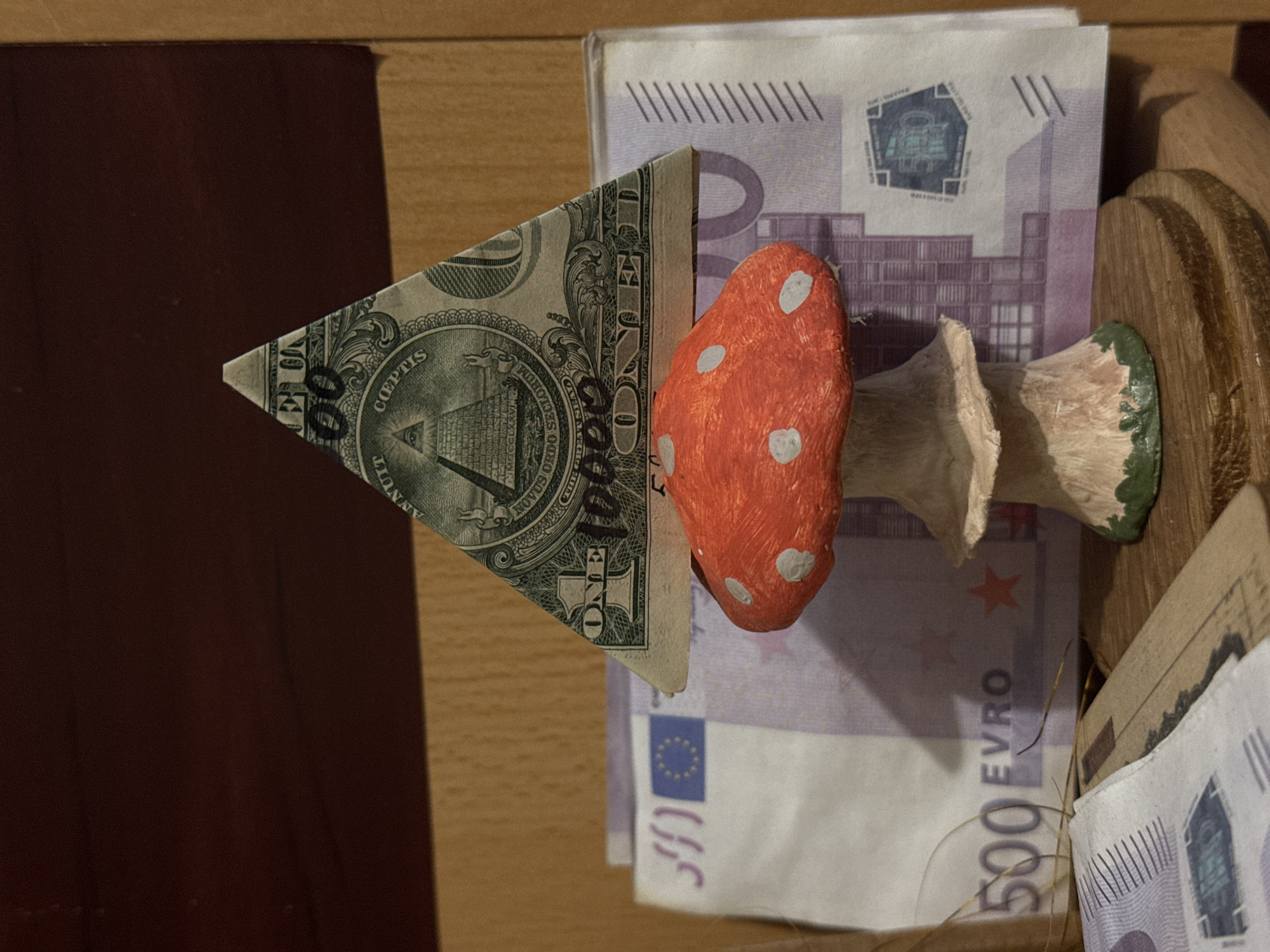 The image shows a still life arrangement of currency and decorative objects. A folded US one-dollar bill forms a triangle, displaying the pyramid and "NOVUS ORDO SECLORUM" on the reverse. Behind it is a stack of Euro banknotes, with a prominent 500 Euro note visible. A decorative mushroom, painted red with white spots, sits on top of the bills. The scene appears to be inside a wooden container. The location of Ново-Никольское, Russia is not identifiable from this image.
