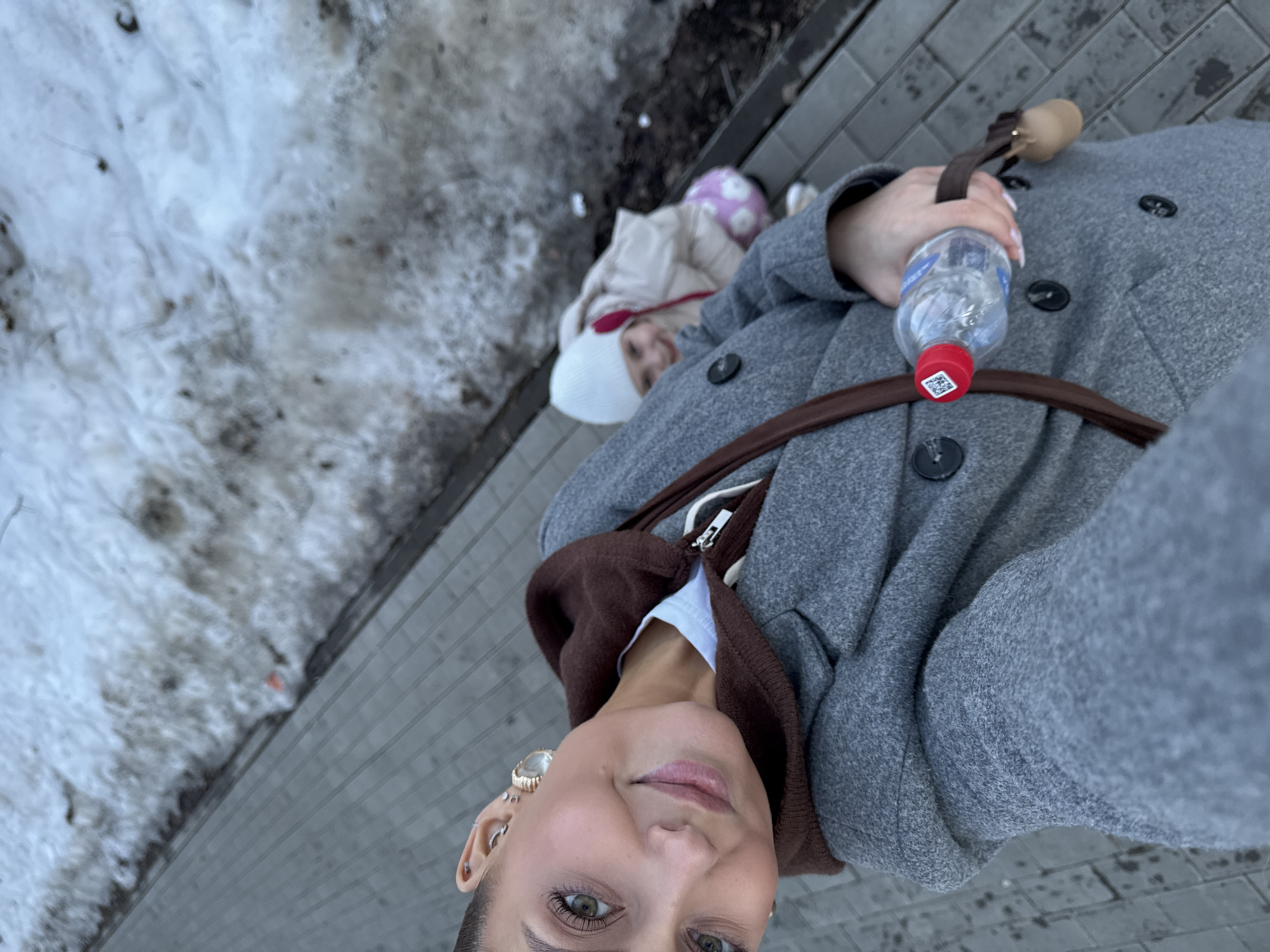 The image depicts an outdoor scene in Chaikovsky, Russia. The primary subject is an adult female, positioned in a close-up selfie-style view, smiling and looking directly at the camera. She has green eyes, dark hair pulled back, and multiple ear piercings, including a prominent gold-colored hoop and a large, clear circular earring. She is wearing a grey wool-blend coat over a brown hooded sweatshirt, with a white garment visible underneath. A brown strap, likely from a shoulder bag, is draped across her. Her right hand, with light-colored manicured nails, holds a clear plastic water bottle with a red cap featuring a white sticker with a black QR code.
In the background, to the right and slightly behind the adult, a young child is partially visible, appearing to be female, crouching or sitting on the ground. The child wears a white knit beanie, a light-colored (possibly beige) padded winter jacket, and pink pants with a white circular or floral pattern, smiling towards the camera.
The ground in the foreground consists of a grey paved sidewalk composed of rectangular pavers. To the right and further back, adjacent to the sidewalk, is an area covered with dirty, melting snow mixed with exposed soil and dark debris, indicative of late winter or early spring conditions.