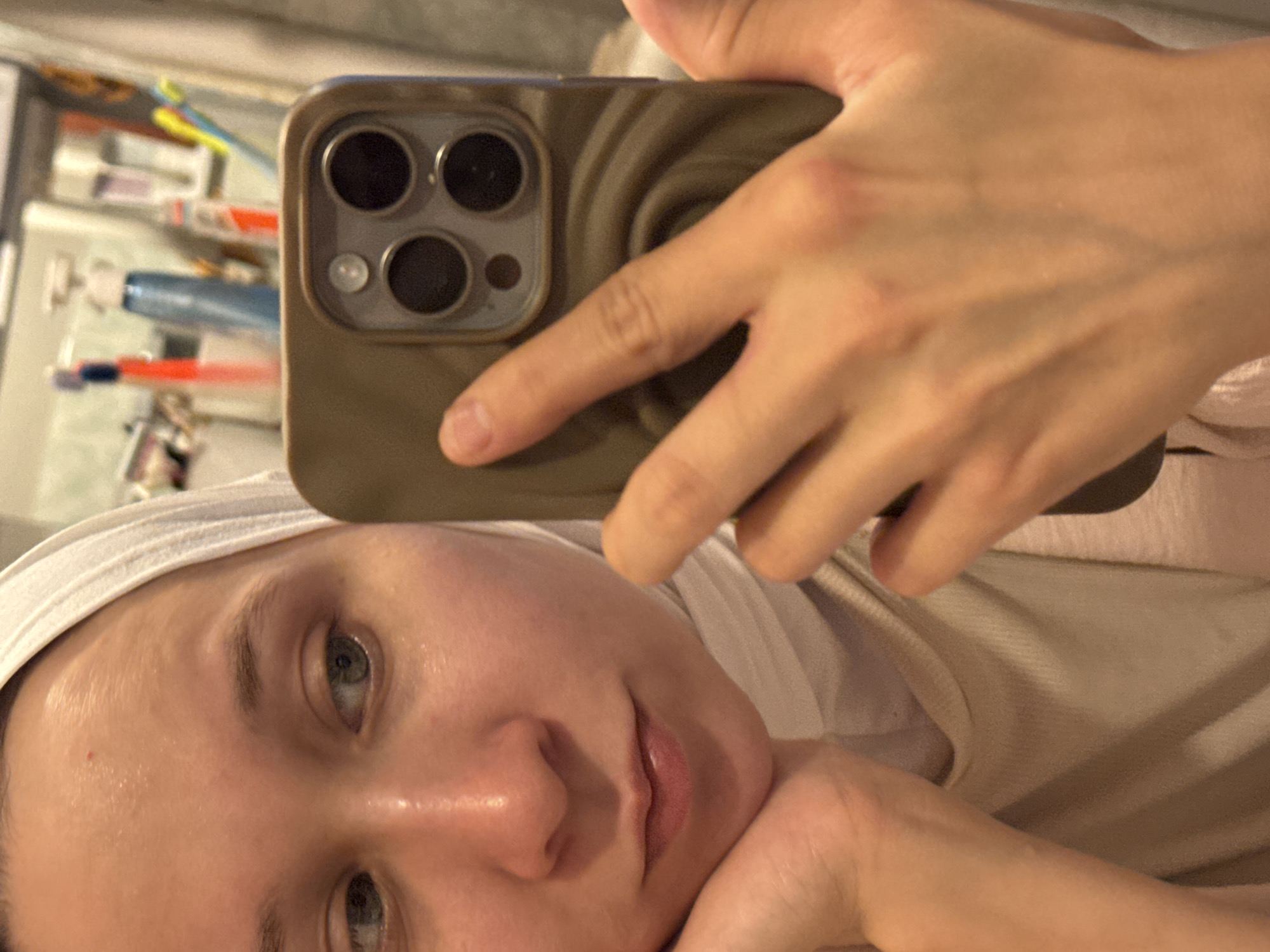 A person is holding a smartphone up to take a selfie. The phone is tan or brown, and the person's hand obscures much of the phone's back. The person appears to be a light-skinned female with blue eyes. She is wearing a white head covering, possibly a towel or headband, and a beige top. The background is blurry and shows a bathroom counter with various toiletries. It is not possible to verify the location of the image based on the current visual information.