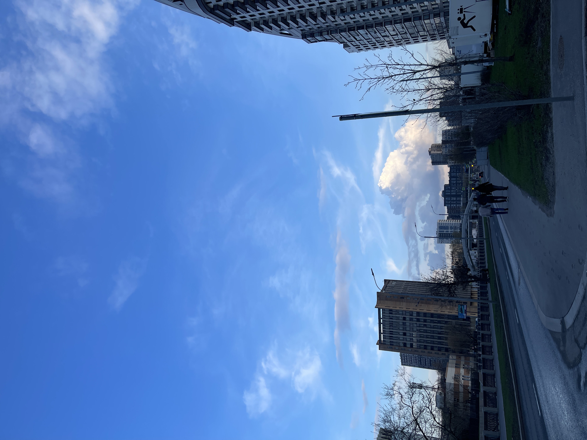 The image is a street-level, vertically-oriented photograph taken outdoors in Moscow, Russia. The viewpoint looks up and along a street lined with modern buildings. The sky is mostly clear blue with wispy white clouds. At the top of the frame is a tall, light-colored building. On the right side of the photo, a sidewalk runs alongside a wet asphalt road. Two people are walking along the sidewalk in the middle distance. The street features streetlights and greenery separating the sidewalk from the road. The image captures a slice of city life under a bright sky in Moscow.
