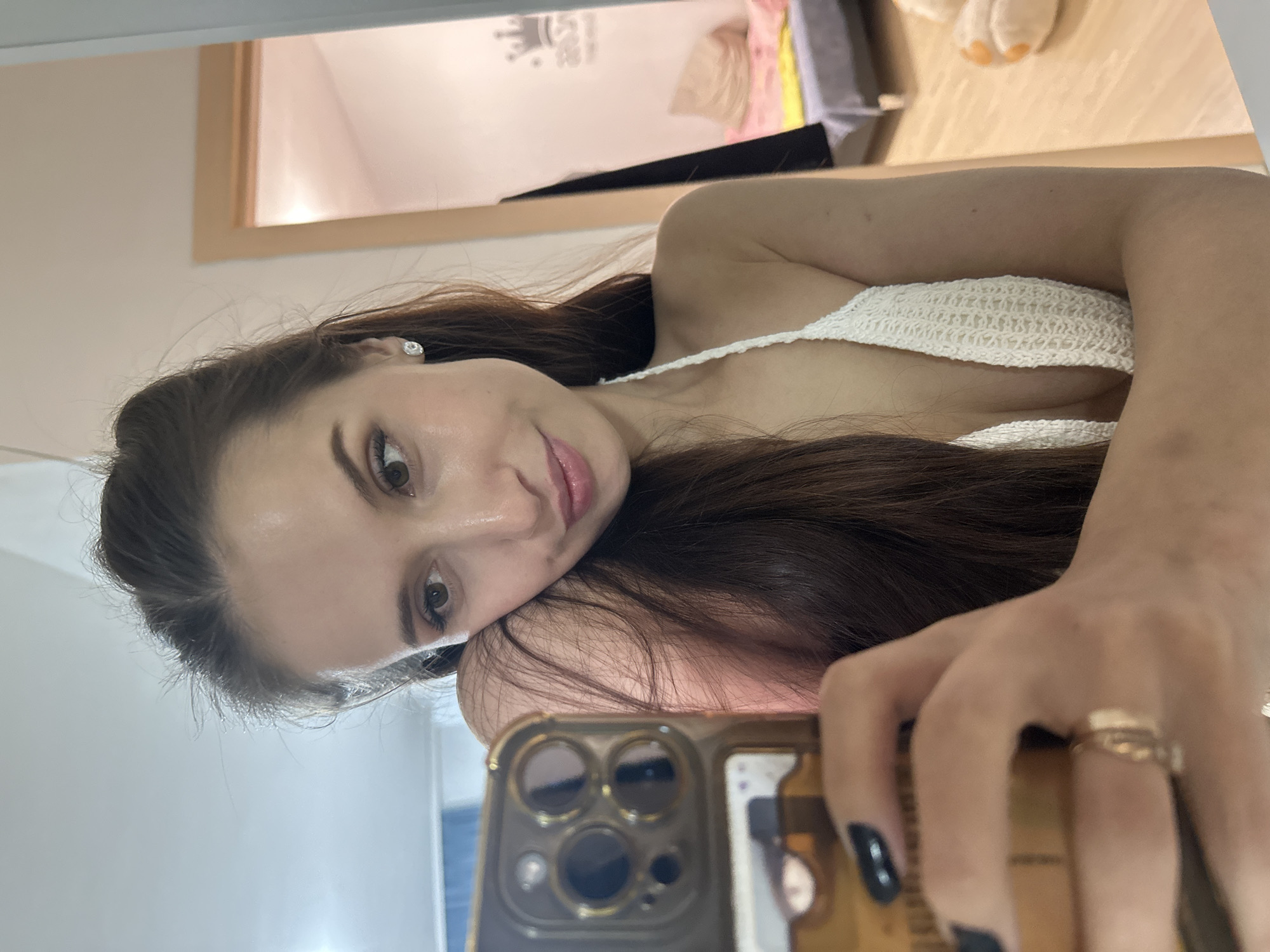 A woman with long brown hair is taking a selfie with a gold-colored smartphone. She is wearing a white crocheted top. A gold ring is visible on her finger. The background includes a glimpse of a room with a framed mirror, and various items suggesting a bedroom or living space are visible, including bedding, a stuffed animal, and wall decor. The location is not discernible from the image.