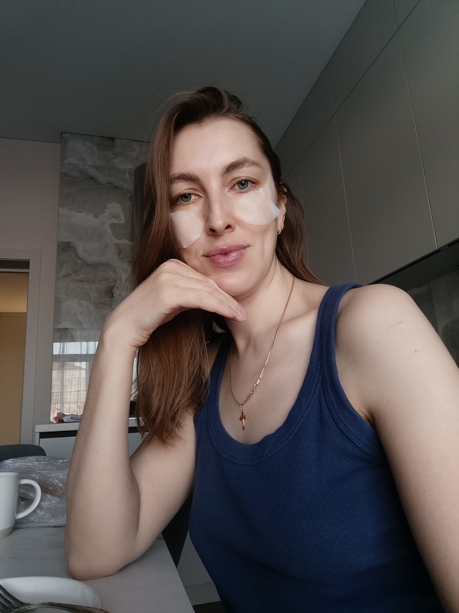 A woman sits at a table. She has fair skin, brown hair, and blue eyes. There are white patches under her eyes. She wears a dark blue ribbed tank top and a gold chain with a cross. Her left arm is propped up, supporting her head. A white mug wrapped in clear plastic sits on the table to her left. A plate and fork are also visible on the table. Behind her is a wall with a light-colored marble-like pattern and a door leading to a room with yellow walls. To her right is a gray wall with visible lines. The photograph seems to be taken indoors in a modern kitchen or dining area. The location is Chaikovsky, Russia.