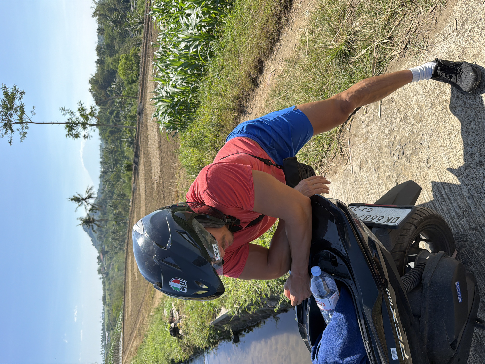 A person wearing a red t-shirt, blue shorts, white socks, and black sneakers is leaning over a black scooter. They are also wearing a black helmet with an AGV sticker. The person appears to be resting or taking a break. A bottle of Evian water is visible on the scooter.
The scene is outdoors, with a rural landscape in the background. There are fields, some with dry stalks and others with green crops, possibly corn. Trees and palm trees dot the landscape, and a hill or mountain is visible in the distance. The sky is clear and blue, suggesting a sunny day. The lighting indicates it is daytime, likely late morning or early afternoon. The environment appears warm and tropical, consistent with the provided location context of Dampit, Indonesia.
The scooter has a license plate with text that reads "DK 6687 KX" and "G3 2J". There is also text on the scooter that says "PCX 160". The overall mood is one of a pause during a journey or activity in a warm, outdoor environment.