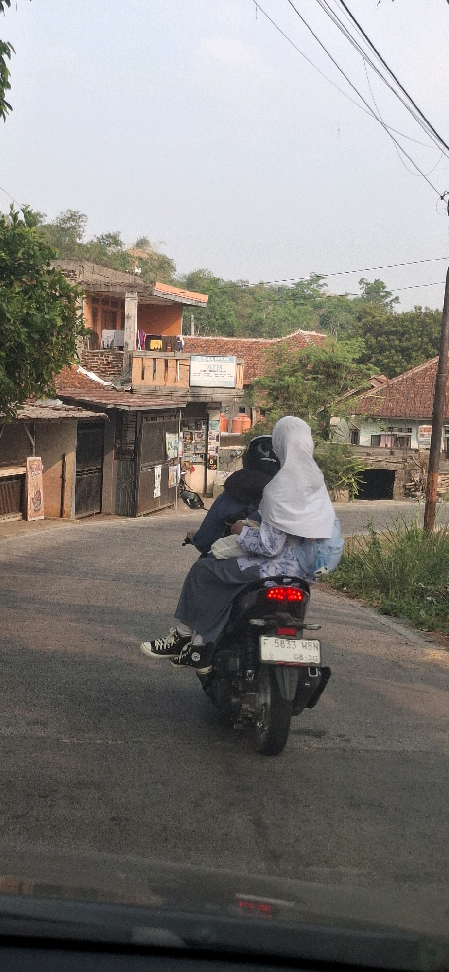 The image shows two individuals riding a motorcycle on a paved road in Ciharashas, Indonesia. The rider, wearing a dark jacket and helmet, is partially obscured. The passenger, wearing a white hijab and a blue and white patterned top with a dark skirt, is seated behind the rider. Both individuals appear to be wearing school uniforms. The motorcycle has a license plate that reads "F 5833 HBN". The environment features buildings with red tiled roofs, green trees, and overhead power lines. One building has signage that includes "ATM". The road surface is asphalt. The sky is overcast and bright. The perspective is from behind and slightly to the side of the motorcycle.