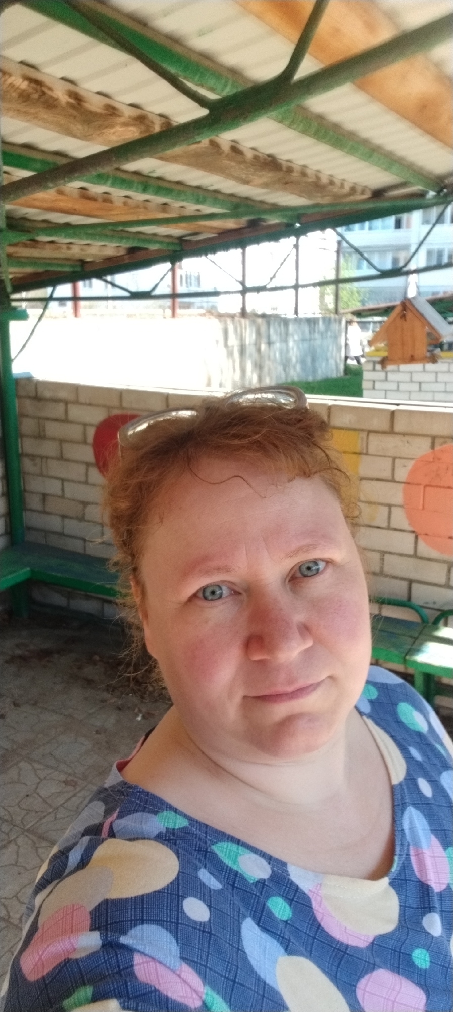 The image depicts a red-haired woman with blue eyes, wearing glasses perched on her head and a blue and multicolored patterned top. She is indoors, under a structure with a corrugated metal roof supported by green beams. Behind her is a low brick wall with painted circles on it, as well as a small wooden birdhouse mounted on the wall. In the background, a building can be seen in the distance.
