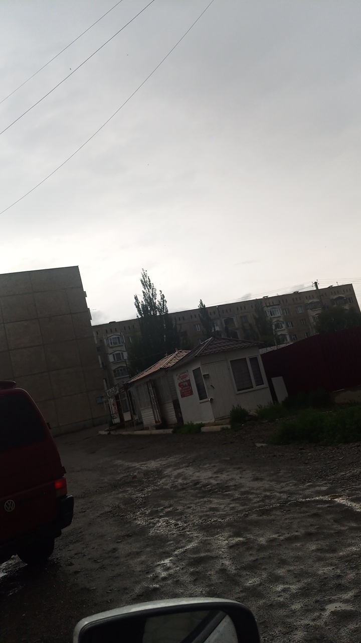 The image depicts an outdoor scene in Balykchy, Kyrgyzstan, likely on an overcast day. In the foreground, the rear of a dark red van is visible on the left. The ground appears to be a wet, rough, unpaved surface with puddles and uneven textures. In the middle ground, there is a small, white, kiosk-like structure with a reddish-brown roof, featuring windows and a sign with red lettering. Behind the kiosk, several tall, slender trees stand in front of a large, multi-story apartment building. This building has a distinct architectural style with rows of dark rectangular openings across its facade, suggesting a Soviet-era construction. To the far left of the image is a portion of a large, gray, Brutalist-style building. A dark red fence is visible to the right of the apartment building. Power lines are present in the sky. The overall environment suggests a residential area in a city.