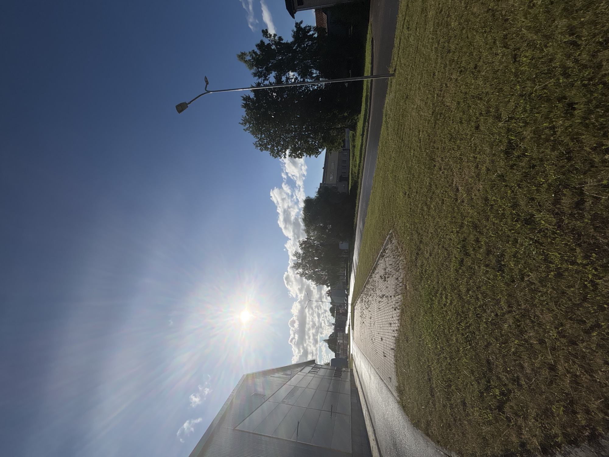 The image depicts a street scene in Sauleskalns, Latvia, under a bright, sunny sky with scattered clouds. The foreground features a well-maintained lawn bordering a paved walkway and a road. A modern building, possibly an industrial or commercial facility, occupies the left side of the frame, its metallic facade reflecting the sunlight. In the middle ground, a solitary lamppost stands near a cluster of mature trees. The background reveals more buildings and foliage, suggesting a developed urban or suburban area. The sun is intensely bright, casting lens flares and creating a radiant effect across the sky, indicating it is likely midday. No people are visible in the scene.