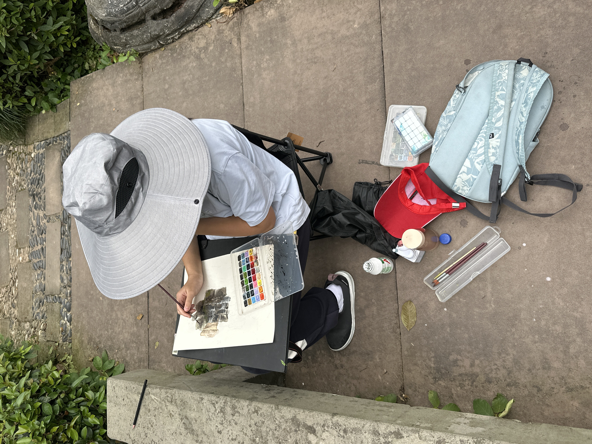 A person wearing a gray sun hat and a light-colored t-shirt is sitting on a black folding chair outdoors, painting a watercolor on a white sheet of paper placed on a black surface. The person is holding a thin paintbrush in their right hand. To the right of the person, on the ground, are a light blue backpack, a red baseball cap, a clear plastic box with watercolor paints, a clear plastic container holding paintbrushes, and a small white bottle. The ground is a mixture of concrete and paving stones. Green plants are visible on the left side of the frame. The location is in the city of Liangjiang, China.