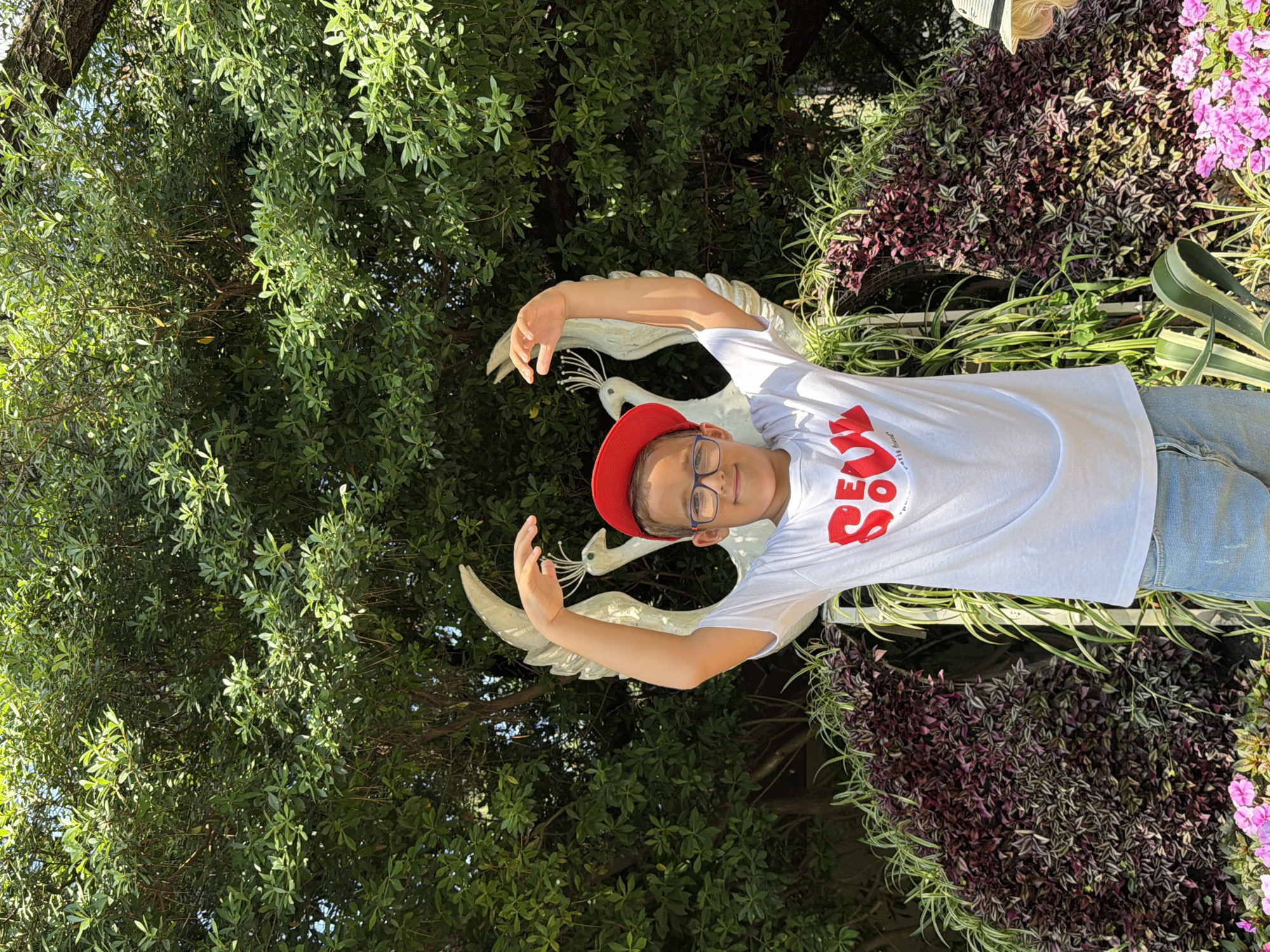 A young boy wearing glasses, a red baseball cap, and a white t-shirt with red lettering that reads "SEOUL" and "paris friend" stands in front of a lush green tree. He has his arms raised above his head, with his hands open. Behind him, a white sculpture of two stylized birds with large wings is visible. The boy is positioned in the center of the frame, and the background consists of dense foliage. The lower portion of the image shows a variety of plants, including deep purple and green leafy plants and some bright pink flowers. The scene appears to be outdoors, possibly in a garden or park. The location is identified as Sochi, Russia.