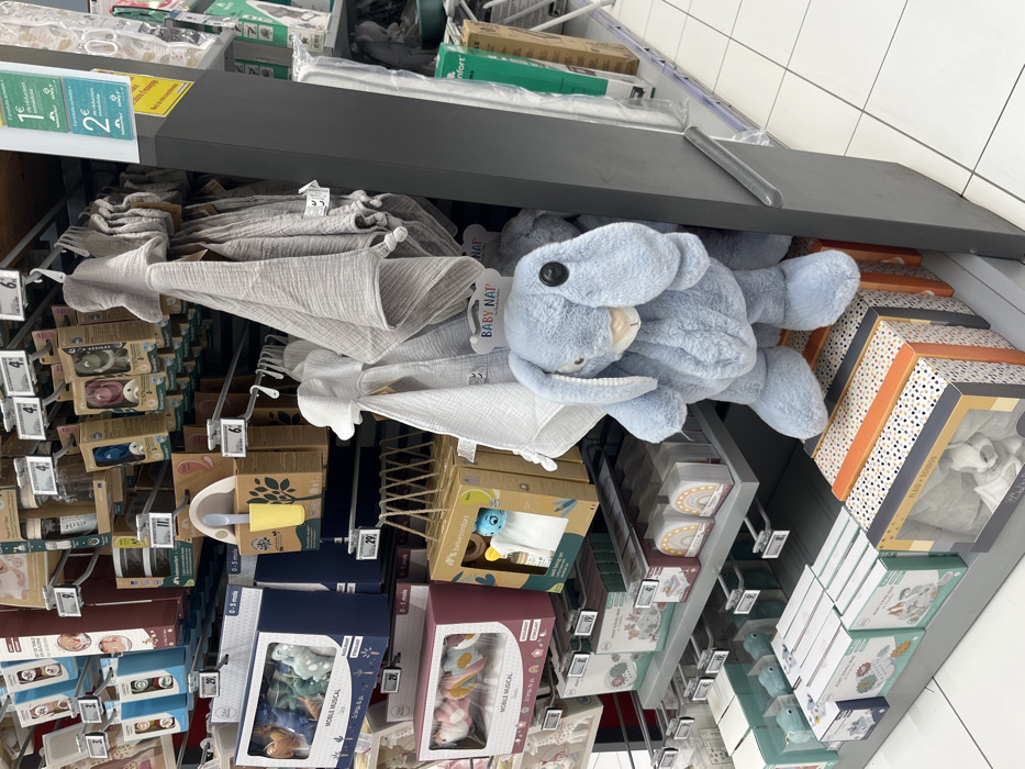 The image shows a section of a retail store in Saint-Brice-Courcelles, France, located at latitude: 49.283, longitude: 3.999. The store aisle features shelves stocked with various baby products. On the left side, there are several boxes of baby toys, including musical mobiles and plush toys. On the right side, there is a large display of baby blankets and swaddles. In the center, a large, blue plush bunny with a button eye sits on a lower shelf, partially obscured by a grey blanket. Below the bunny, there are several boxes of baby clothes, including a box labeled "PLAID DOUDOU" and a box with a picture of a baby wearing a white onesie. The overall scene depicts a typical retail environment showcasing baby products.
