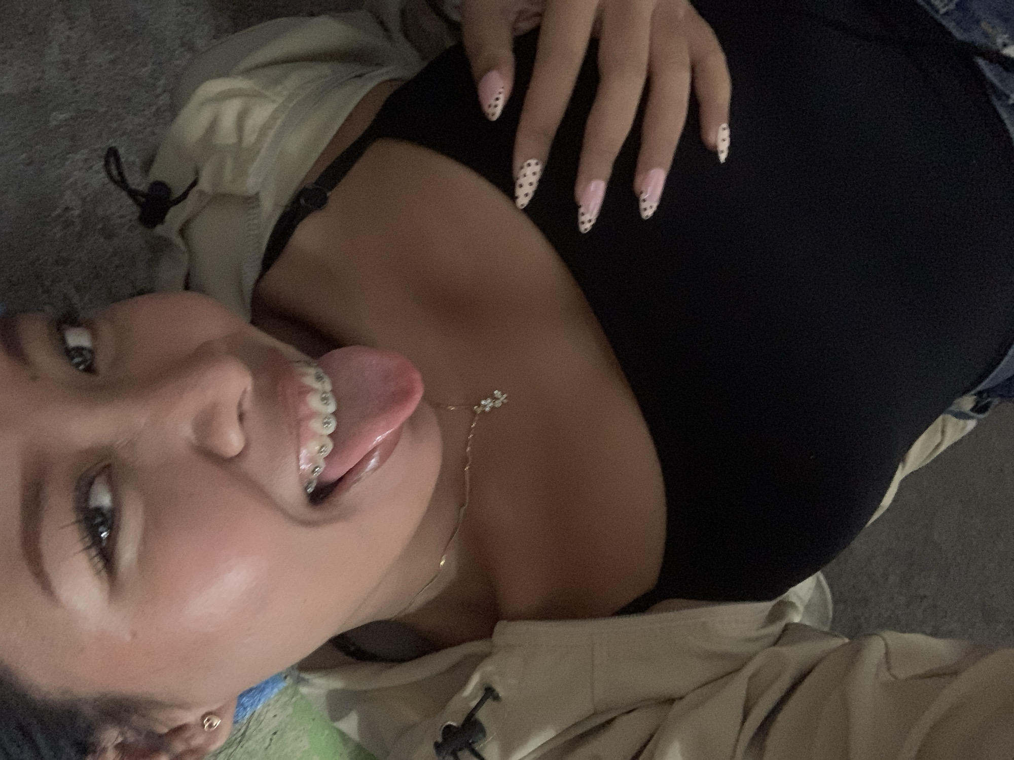 A young woman with braces is lying down and sticking out her tongue. She is wearing a black tank top and a tan jacket. Her nails are painted pink with black polka dots. She is also wearing a gold necklace with a small pendant. The background appears to be an indoor setting, possibly a home, with a textured floor. There is no indication of the location being in Tamanbaru, Indonesia, or any specific city.