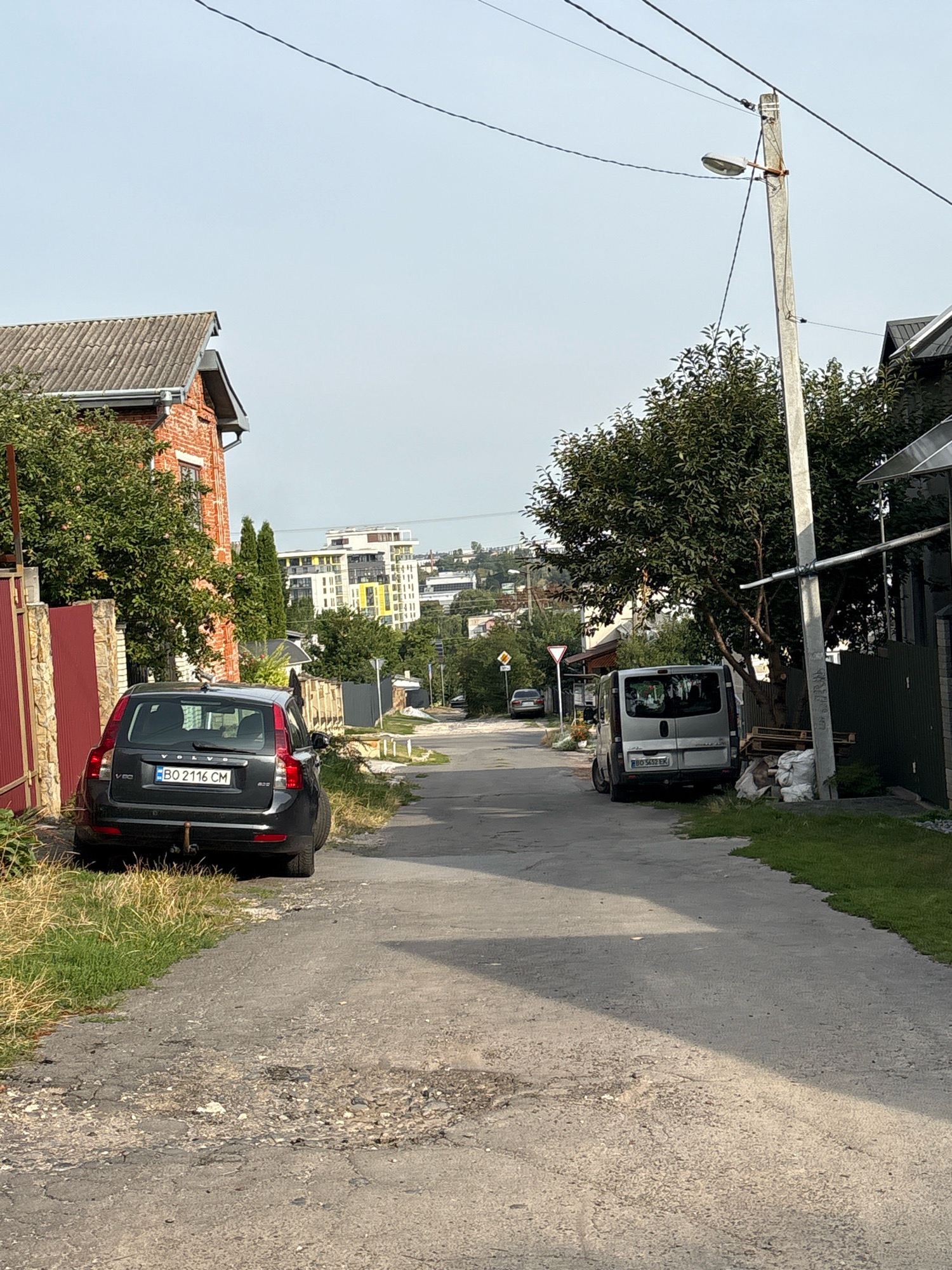 The image depicts a narrow asphalt road in Ternopil, Ukraine, lined with vegetation and residential structures. In the foreground, a dark grey Volvo station wagon is parked on the left side of the road. Further down the road, a silver minivan is parked on the right. Several trees are visible, with dense foliage. In the background, modern apartment buildings stand out against the sky. The road surface is uneven, with visible cracks and patches. The sky is clear and pale. The scene appears to be a quiet residential street, likely in the suburbs of Ternopil.