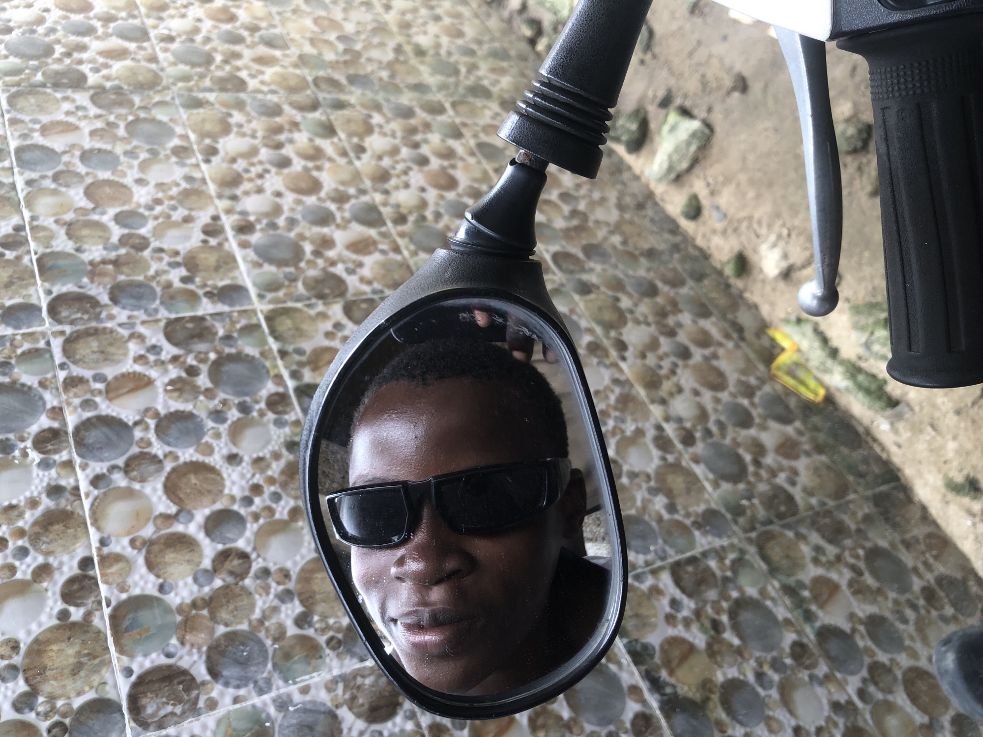 A close-up, eye-level shot shows a reflection of a young Black person wearing dark sunglasses in the mirror of a motorcycle. The person's head is tilted back, and their eyes are closed, with their face illuminated by sunlight. The mirror itself is black with a silver stem, attached to a black handlebar with ribbed grips and a silver brake lever. The background is a wall covered in mosaic tiles with circular patterns in shades of brown, gray, and beige. The ground in front of the wall appears to be sand or dirt with scattered rocks and some yellow and green debris. The scene is set outdoors, likely on a street or pathway, possibly in Ajah, Nigeria, given the context.