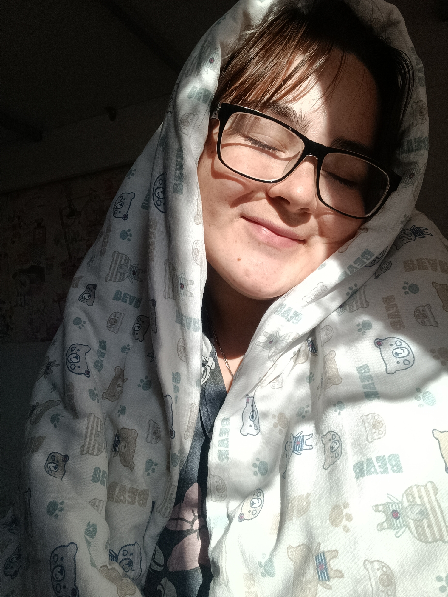 A young person with brown hair and glasses is wrapped in a white blanket patterned with cartoon bears and the word "BEAR". They are smiling with their eyes closed. Sunlight illuminates the right side of their face and the blanket. The background shows patterned wallpaper. The location, Noviy, Russia, is not visually verifiable.