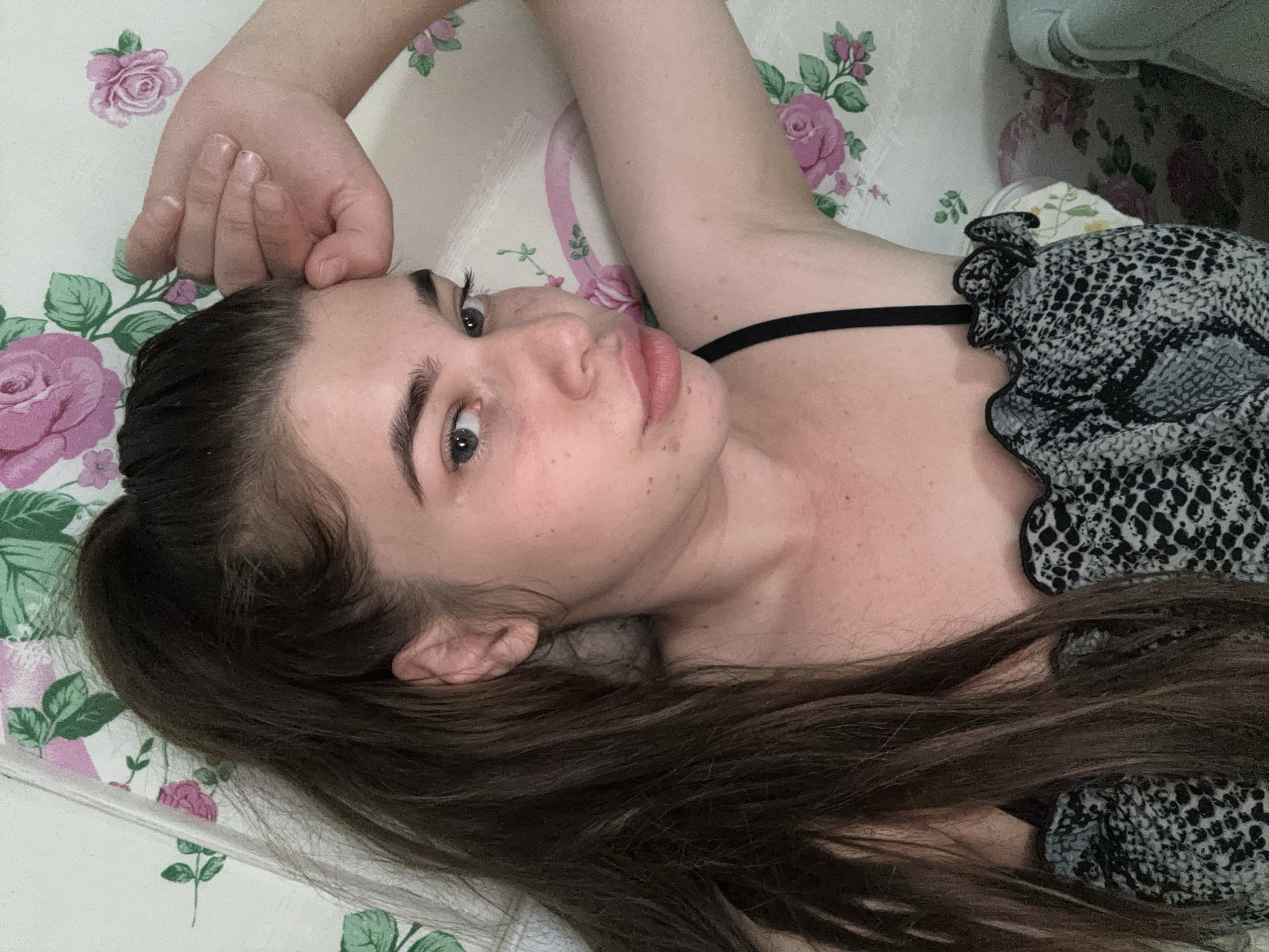 A young woman with long, dark brown hair pulled into a high ponytail is lying on her back. She is looking up at the camera with a neutral expression, her right hand resting on her forehead. She is wearing a black camisole with a patterned grey and white fabric detail at the neckline. The background is a floral-patterned sheet. The image does not provide any information about the location in the city of Мещёры, Russia.