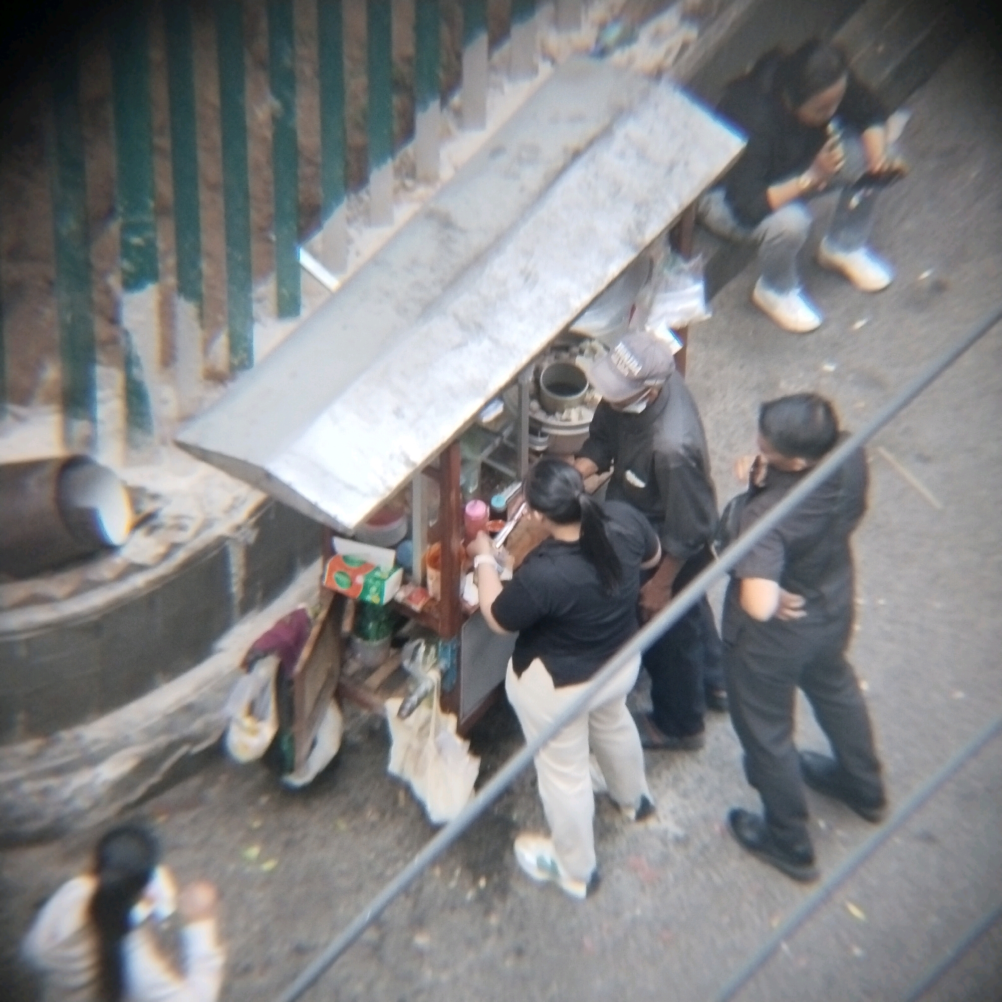 In Surabaya, Indonesia, at an outdoor food stall, a group of people are engaged in various activities. A man wearing a black shirt and a cap stands by the stall, while a woman in a black shirt and light-colored pants prepares food. To the left, another person in a light-colored shirt and dark pants is seated, looking at their phone. Further back, a person in a black shirt and dark pants is also present. The stall itself is covered with a silver-colored canopy and contains various containers and bottles, suggesting food preparation. The ground is paved, and a metal railing runs diagonally across the image. Green bars are visible in the background.