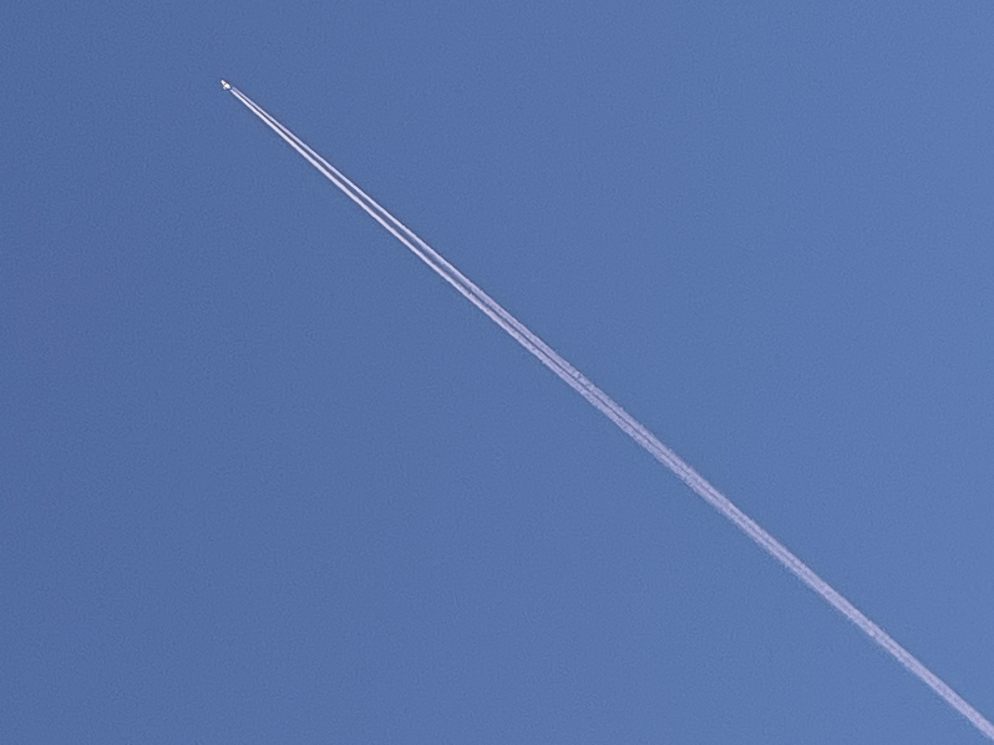 The image shows a jet plane high in the sky, leaving a white contrail. The sky is clear and blue. The location of the image is not discernible from the content of the picture. The city of Chaikovsky, Russia, has no discernable relevance to the image.