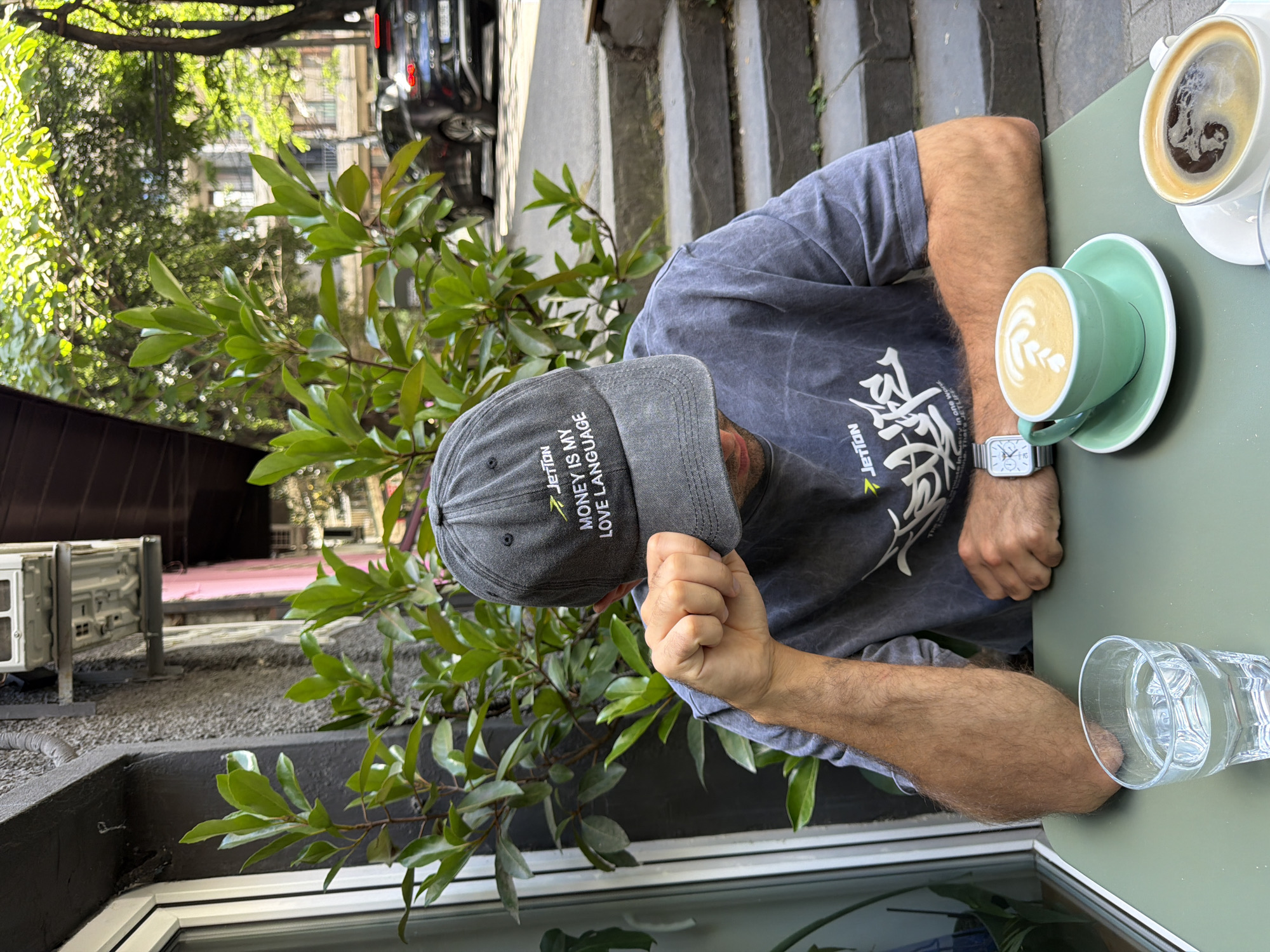 A person, seen from the chest up, wearing a grey t-shirt and a grey baseball cap, is sitting at a table outdoors. The person's hand is resting on the table, and they are looking down. On the table, there are two cups of coffee and a glass of water. The coffee cup in the foreground is light green and contains a latte with latte art. The other coffee cup is white and contains a darker coffee. The person is wearing a silver watch on their left wrist. The background shows greenery and a building with parked cars, suggesting an urban setting. The baseball cap has text that reads "JETTON MONEY IS MY LANGUAGE". The t-shirt also has some text and graphics on it, including "JETTON". The scene appears to be in a cafe or an outdoor seating area. The lighting suggests it is daytime, likely morning or afternoon, with sunlight filtering through the trees. The overall mood is casual and relaxed.