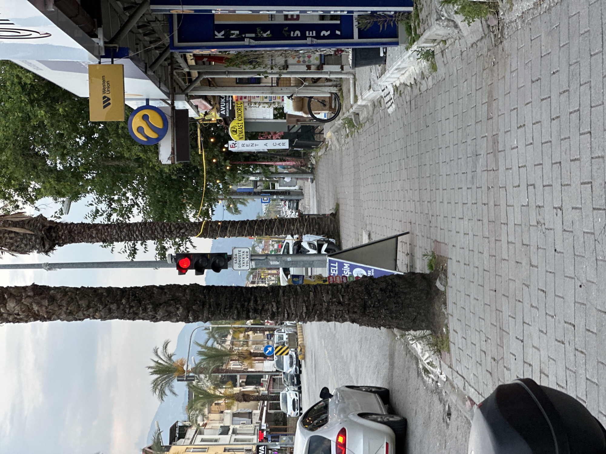 The image shows a street scene in Fethiye, Turkey. The view is from a sidewalk paved with gray brick leading into the distance toward mountains. Palm trees line the street, partially obscuring the view. A traffic light is visible, showing a red light. A sign instructs drivers to yield to pedestrians when turning right. Street signs for businesses like Western Union, Avuka, and Rent a Car are visible. Vehicles are parked along the road, including a white car in the foreground and other vehicles further down the street.