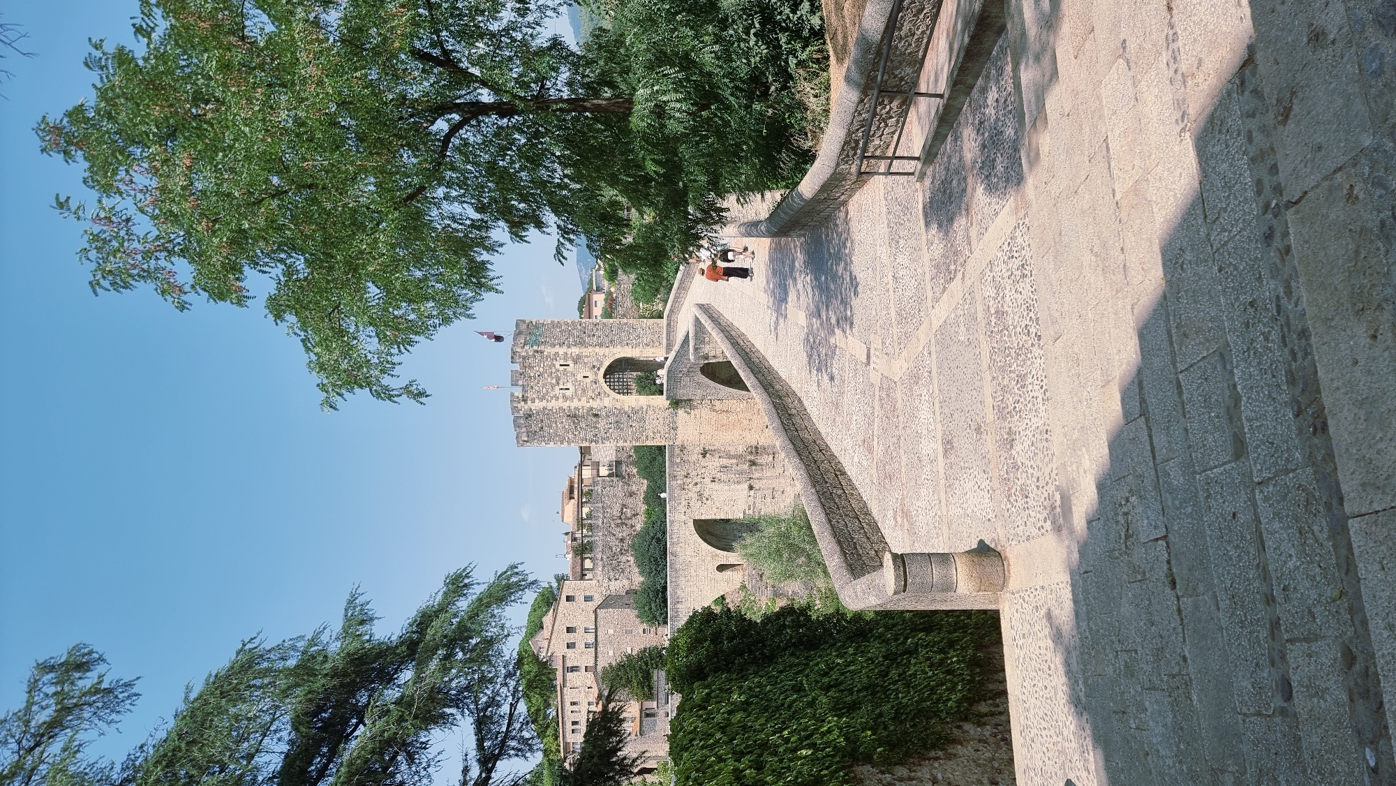 The image shows the Pont Fortificat (Fortified Bridge) in Besalú, Spain, a medieval stone bridge with a fortified gatehouse at its center. The bridge spans a dry riverbed or canal. The bridge and gatehouse are constructed of stone blocks, with arched sections and a square tower topped with battlements. Two people are walking across the bridge, one in an orange shirt and the other in a white shirt. To the left of the bridge, a stone wall is covered in dense green ivy. The background reveals more stone buildings and walls, characteristic of the old town of Besalú. Trees with green foliage frame the scene on the left and right, with a clear blue sky overhead. The foreground shows a stone stairway leading down towards the bridge.