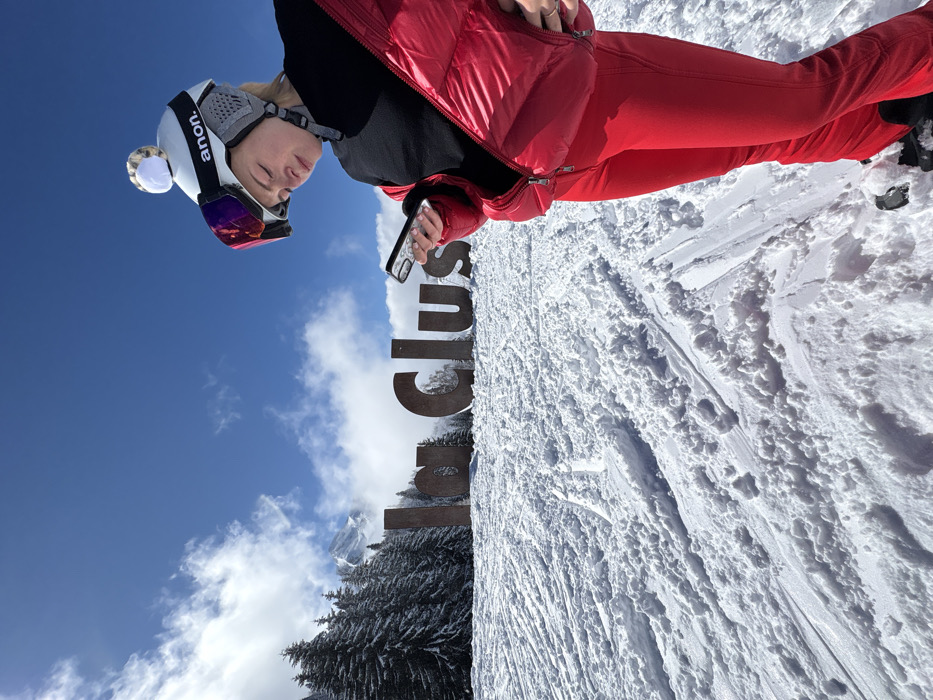 The image depicts a snowy outdoor scene at the La Clusaz ski resort in France. In the foreground, a person, appearing female, stands on a snow-covered slope, partially turned towards the left. This individual wears a white Anon ski helmet adorned with a light-colored pom-pom, ski goggles with mirrored red lenses pushed up onto the helmet, a black long-sleeved top, a shiny red puffer jacket unzipped to the waist, and red ski pants. Black ski boots are visible at the bottom. The person is actively engaged with a black smartphone held horizontally in both hands, looking down at the screen.
In the mid-ground, behind the individual, a large, dark, weathered sign spells out "La Clusaz" in capital letters, embedded in the snow. To the left of the sign and in the background, a dense forest of snow-laden coniferous trees extends up a slope. Distant snow-capped mountains are visible beyond the treeline under a bright blue sky with scattered white clouds. The ground is covered in deep, textured snow, marked by numerous ski tracks and indentations. The sun casts bright light, indicating clear weather.