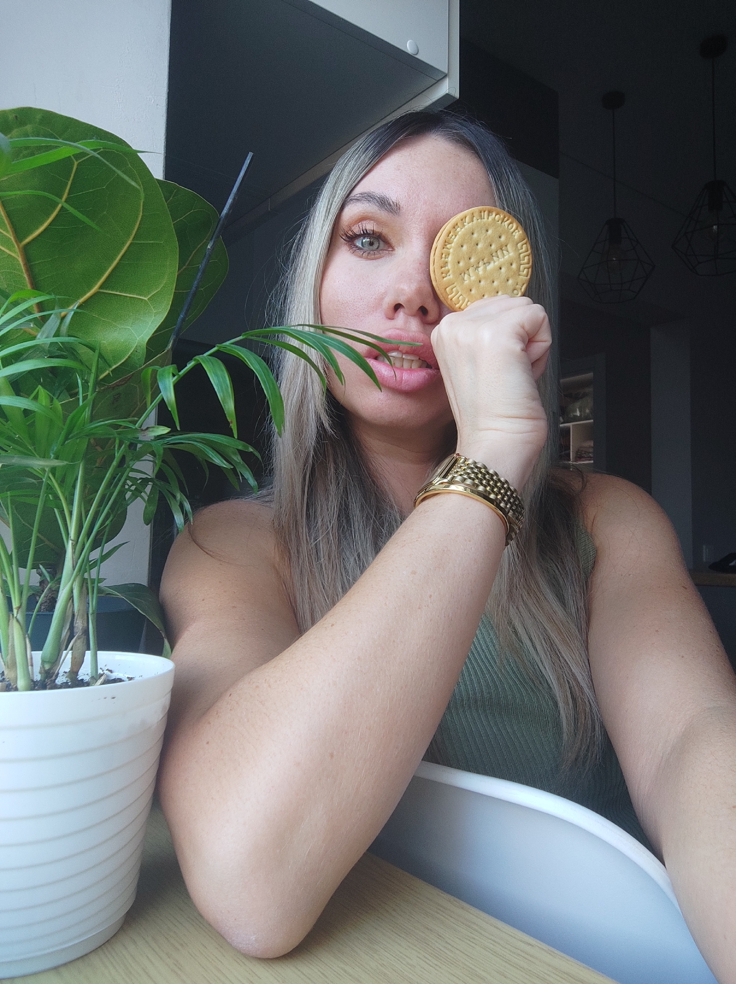 A woman is seated indoors, possibly at a table, holding a round biscuit over her right eye. She wears a green ribbed top and a gold bracelet on her right wrist. A potted plant with green foliage sits to her left. The woman has long, light brown hair with blonde highlights. In the background are light fixtures hanging from the ceiling. The location cannot be confirmed as Votkinsk, Russia from the image alone.