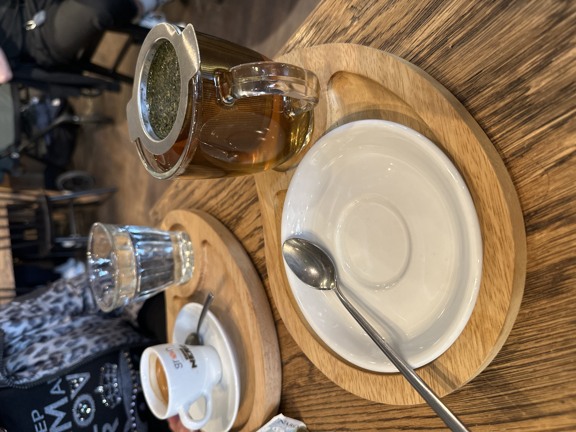 The image captures a cafe scene in Wrocław, Poland, depicting a wooden table with two wooden serving trays. On one tray, there is a glass teapot with a metal infuser filled with tea leaves, containing a light brown liquid, and a white saucer with a metal spoon. The second tray holds a small, white espresso cup with a saucer, a small glass of water, and part of a black purse with a white and gray pattern. The background shows parts of cafe furniture and people, suggesting the image was taken indoors during daylight.