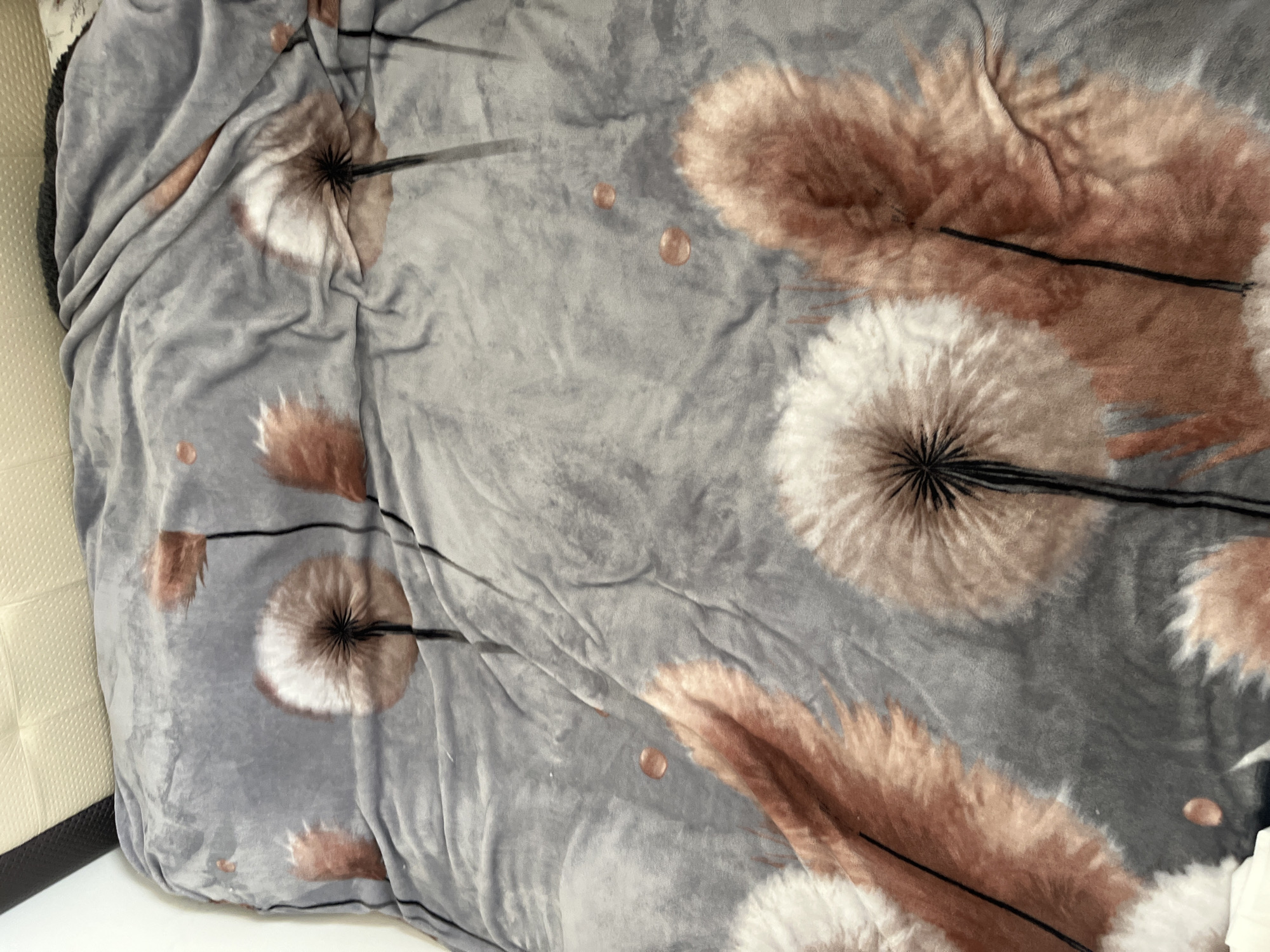 A close-up, high-angle shot shows a grey blanket with a dandelion print spread out on a bed in Onokivtsi, Ukraine. The blanket features large, stylized dandelions in shades of white, brown, and orange, with thin black stems. The fabric is soft and slightly creased, indicating it has been used. A portion of a headboard is visible in the upper left corner, appearing to be upholstered in beige or cream material with a textured pattern. The rest of the bed is covered by the blanket. The scene is indoors, likely in a bedroom, with natural light possibly filtering in from an unseen source. There are no people or animals present in the image. The overall impression is one of domestic comfort.