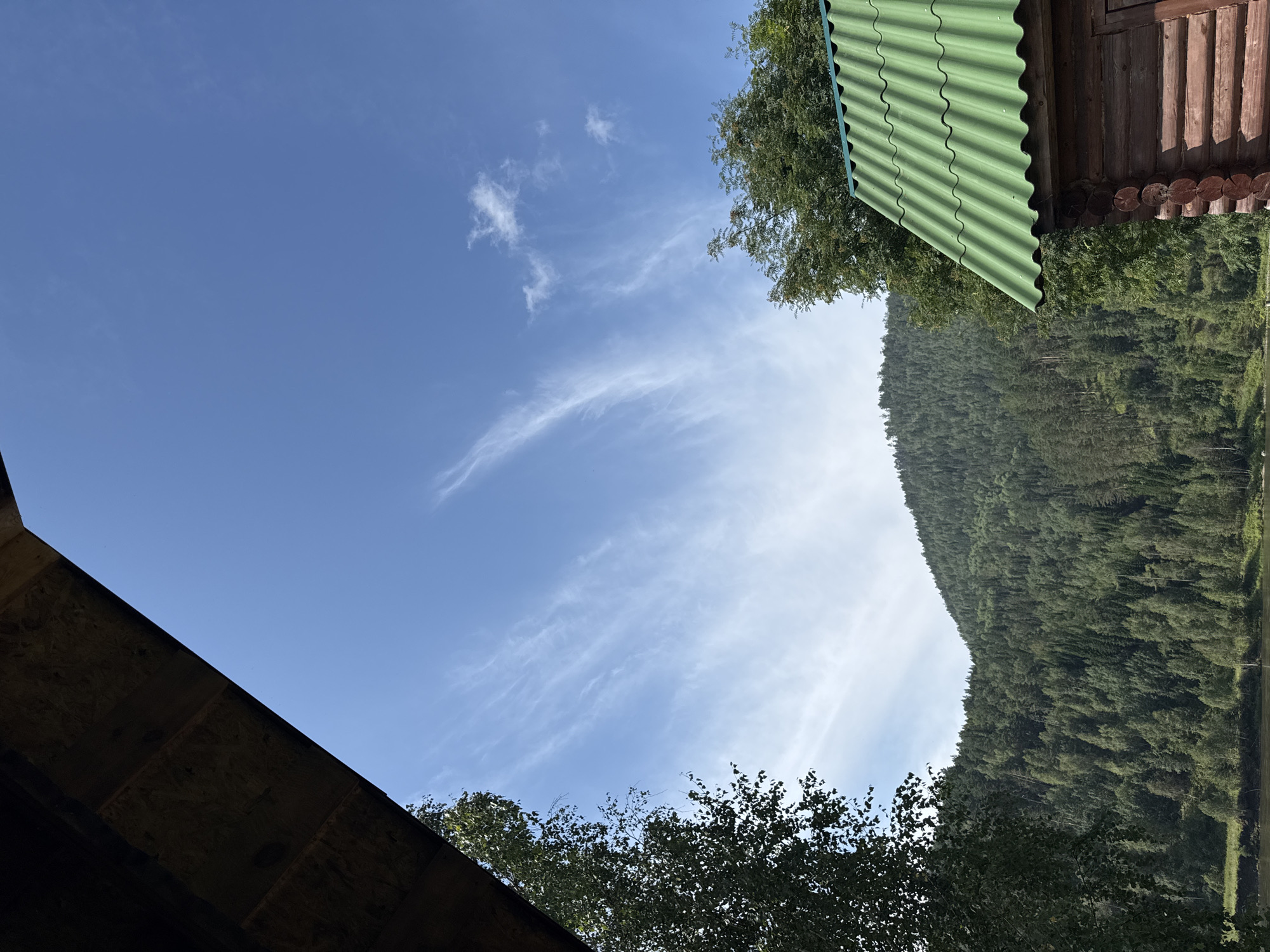 The image captures a tranquil scene in Ust-Mana, Russia, framed by architectural elements and natural landscapes. In the foreground, a wooden structure with a green corrugated roof is partially visible on the right, suggesting a rural dwelling or outbuilding. To the left, the underside of a dark wooden roof overhang creates a natural border.
Beyond the structures, a dense forest covers a rolling hill, displaying a rich tapestry of green from various coniferous and deciduous trees. The forest extends towards the background, where the terrain rises into a mountainous landscape.
The sky above is a vibrant blue, sparsely dotted with wispy white cirrus clouds, indicating fair weather. The overall impression is one of serene natural beauty, likely a view from a balcony, porch, or covered area within a settlement near the forest. There are no people or animals visible in the frame.