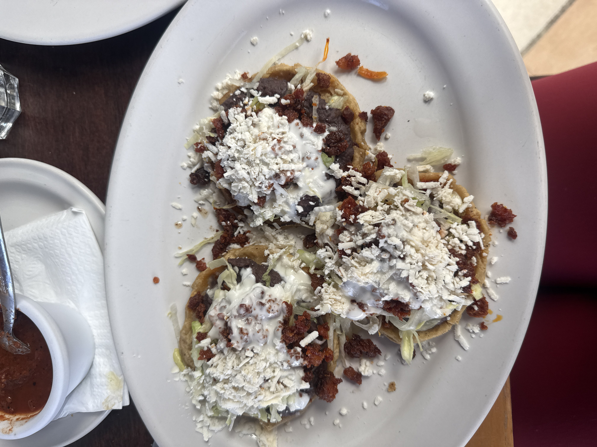 A top-down, medium shot depicts a white plate holding three sopes. The sopes are topped with lettuce, beans, crumbled chorizo, white cream, and a generous amount of crumbled cheese. To the left of the plate is a small white dish filled with red salsa and a spoon. The plate and salsa dish are resting on a dark-colored table. Part of another white plate is visible in the upper left corner of the frame, and a red chair back is visible on the right. The location is Unknown, Unknown.