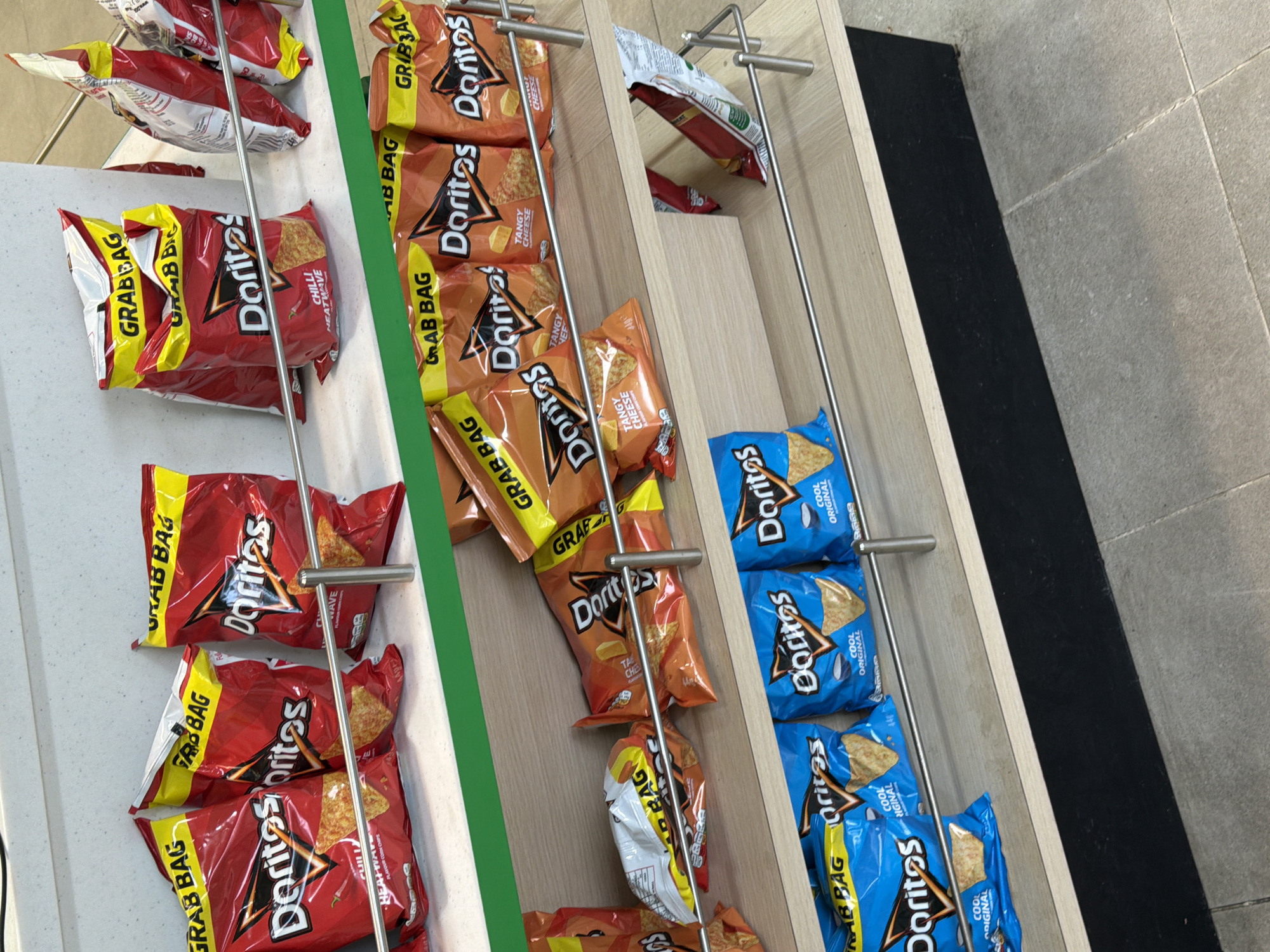 The image shows a display rack in a retail environment, likely a convenience store or gas station, situated in Pity Me, United Kingdom. The rack is organized into three columns and features several rows of "Grab Bag" size Doritos chips. The left column contains Doritos Chili Heatwave. The middle column contains Doritos Tangy Cheese. The right column contains Doritos Cool Original. The chip bags are arranged behind metal bars that keep them in place. The rack itself is constructed of light-colored wood with a green trim piece between the first two chip columns. The base of the rack is adjacent to a section of dark-colored flooring. The background shows tiled flooring. The scene is illuminated with artificial light. There are no people visible in the image.