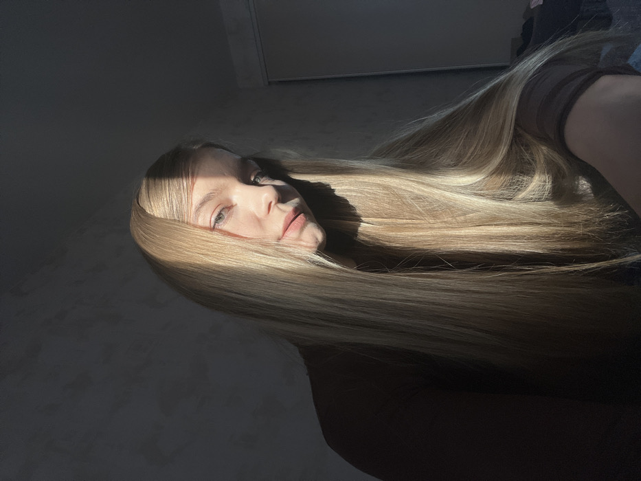 The image depicts a young female individual indoors, located in the city of Zvezda, Russia. She is positioned towards the center-right of the frame, facing generally forward with her head slightly tilted. Her long, straight blonde hair flows over her shoulders and chest. Her eyes are light in color, and her lips are natural. She is wearing a dark brown, long-sleeved top with a subtly sheer texture on the sleeves. Direct sunlight illuminates the right side of her face and hair, casting a prominent shadow across the left side of her face and body. The background features a dark, textured wall. A vertical light-colored panel, consistent with a door or wardrobe, is partially visible on the far right behind her. The environment is dimly lit overall, with a strong light source from the right creating high contrast.