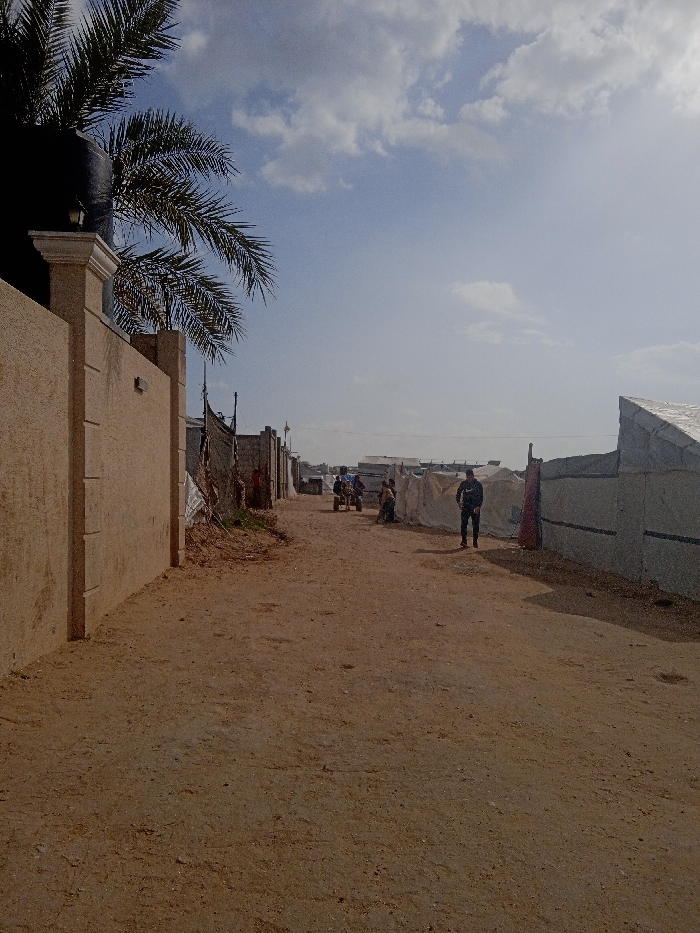 The image depicts a dirt road in Khan Yunis, Palestinian Territory, under a partially cloudy blue sky. On the left, a tall, light-brown concrete wall with a decorative white capstone extends into the midground. Behind the wall, a dark water tank and palm tree fronds are visible. Further down the left side, makeshift structures of various materials line the road. On the right, a series of temporary shelters or tents constructed from white and gray tarpaulin sheets extend along the road. In the midground, a lone male figure in dark clothing stands on the dirt path, facing left. Further down the road, a small group of individuals is present, one seemingly operating a three-wheeled cart or vehicle, and another seated near the makeshift structures. The unpaved road recedes into the background, where additional structures are vaguely visible. Sunlight illuminates the scene, casting shadows.