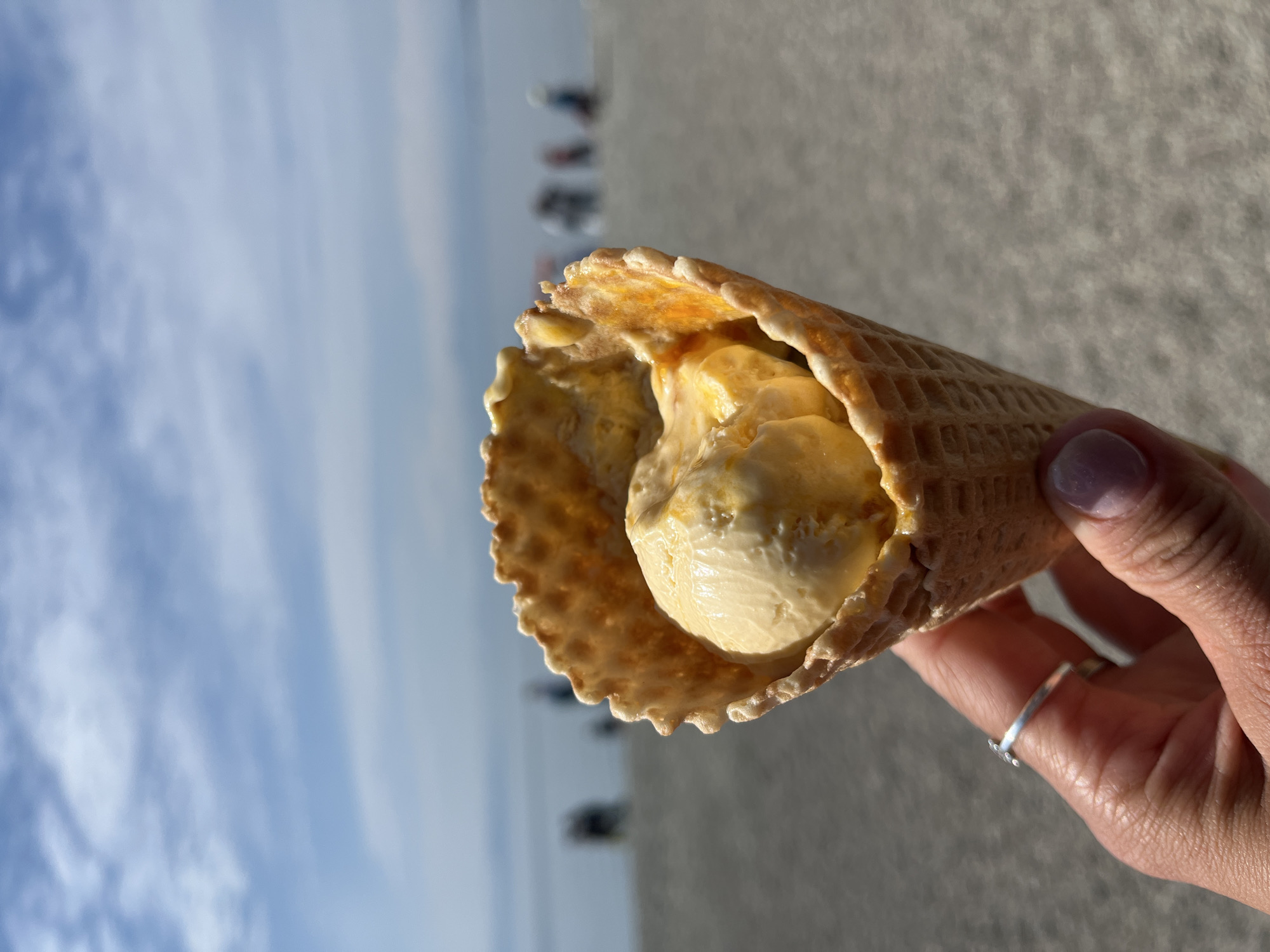 A close-up shot focuses on a waffle cone filled with two scoops of yellow ice cream, held by a person's hand adorned with a silver ring. In the blurred background, a beach scene unfolds with people walking along the shore and the calm, blue ocean stretching towards the horizon under a partly cloudy sky. This image was captured in Petropavlovsk-Kamchatsky, Russia.