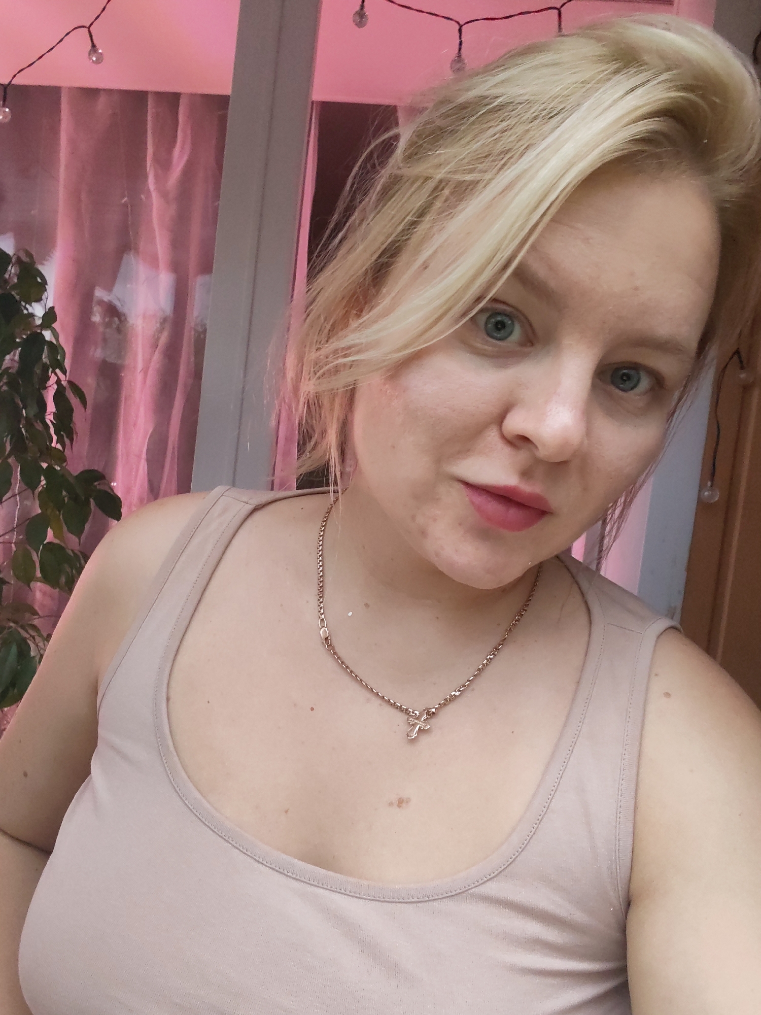 The image shows a young Caucasian woman with blonde hair and blue eyes, wearing a beige tank top and a thin gold chain with a cross pendant. She is looking at the camera with a slight smile. The background is blurred, but appears to be an indoor setting with pink curtains and some greenery. There are also string lights visible in the background. The woman's skin has some light freckles and appears to have a natural tone. The image is taken from a slightly low angle, focusing on the woman's face and upper body.