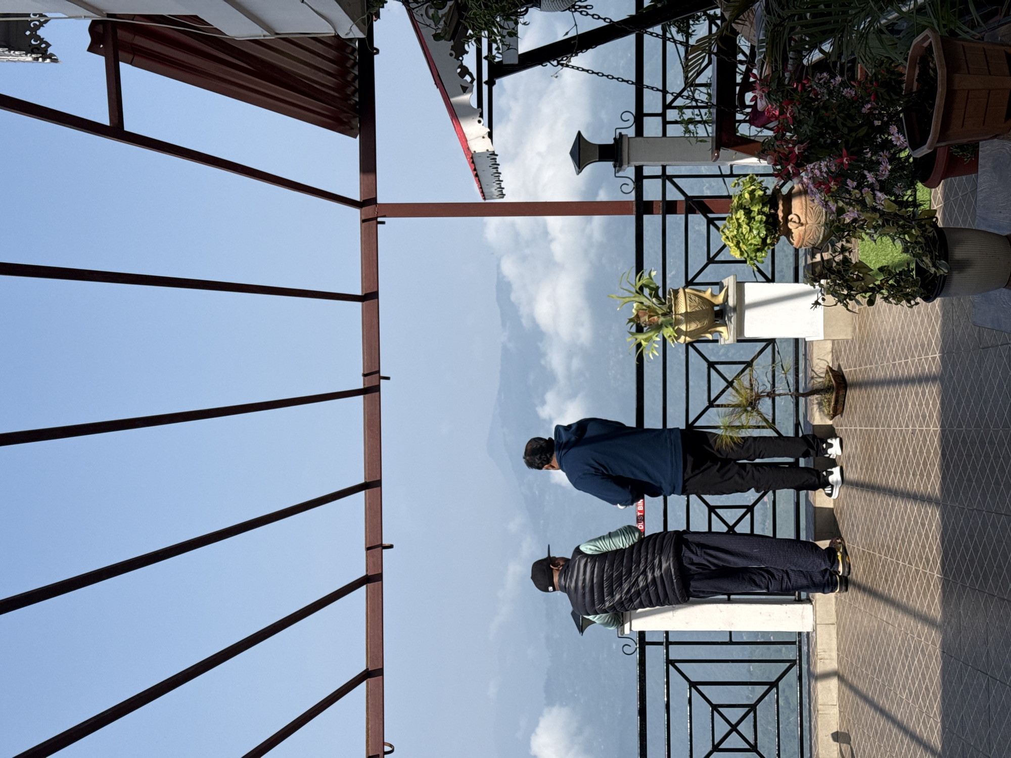 This media file captures a scenic view from a balcony or viewing platform, likely in a mountainous region such as Kurseong, India, under clear daylight conditions.
The main subjects are two men standing at a dark metal railing, looking out towards a vast mountain range partially veiled by clouds. The man in the foreground, facing away from the camera, wears a black puffer vest over a lighter long-sleeved shirt, dark pants, and a black baseball cap. His shoes are dark with a yellow accent. Adjacent to him, the second man wears a dark blue hoodie, black pants, and white and black sneakers. Both appear to be observing the panoramic vista.
The setting is an outdoor area with a patterned tiled floor, reflecting sunlight and casting clear shadows. The railing has a geometric design. Numerous potted plants with vibrant flowers and green foliage decorate the platform, placed on pedestals and directly on the floor. Above, a reddish-brown metal framework, possibly part of a roof or awning structure, spans across the left and top portions of the image.
The background reveals a magnificent mountain landscape stretching into the distance under a bright blue sky with scattered white clouds. The activity taking place is that the two men are admiring the natural scenery, seemingly enjoying a moment of contemplation or observation. The clear skies and bright sunlight suggest pleasant weather. Visible text on a red label near the man in the puffer vest reads "...UST BIN", likely indicating a "DUST BIN".
