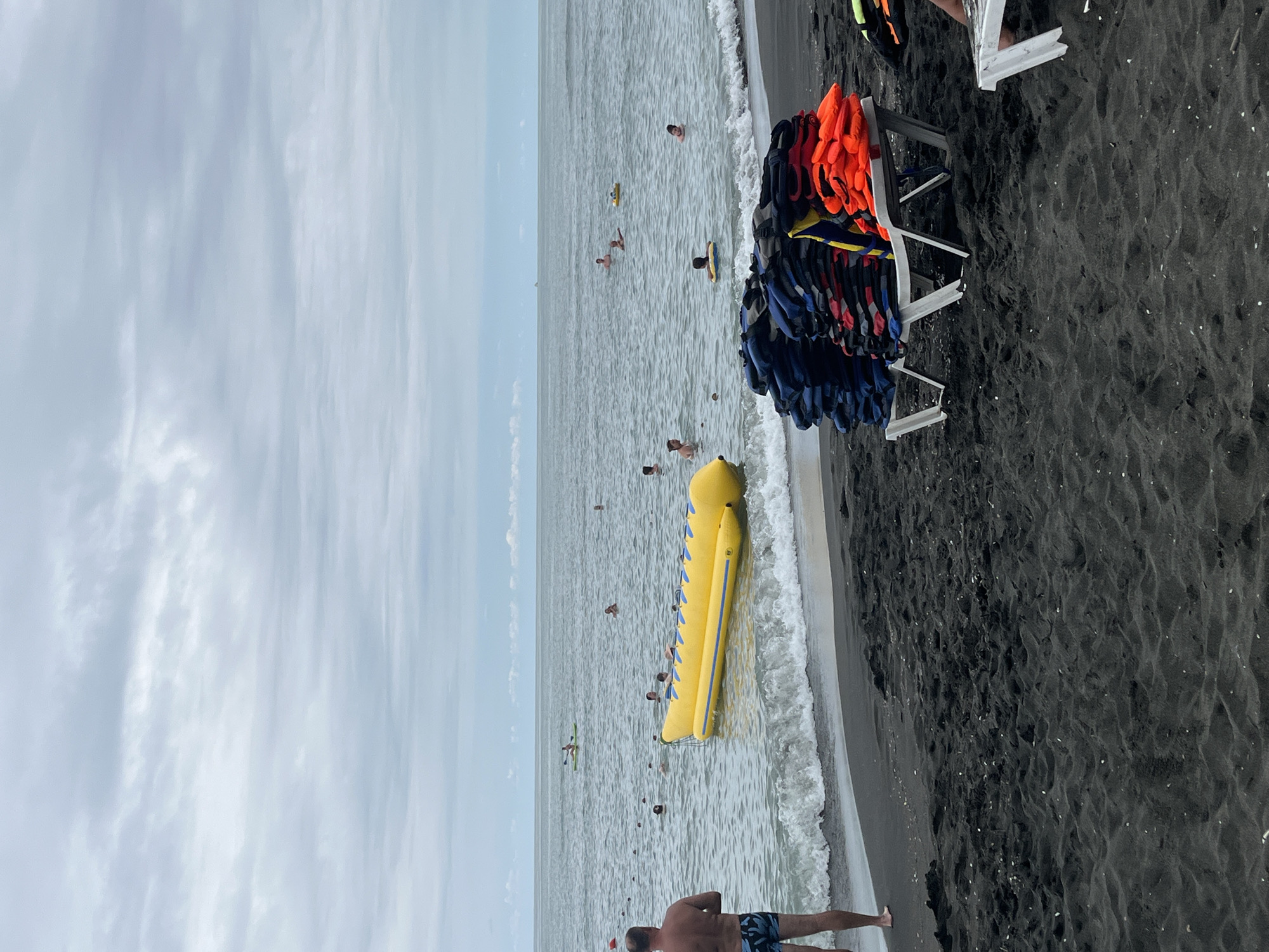 This image shows a view of the Black Sea coast in Ureki, Georgia. The beach has dark sand and the water is calm with small waves. Several people are swimming or relaxing in the water, some with inflatable rings. A large yellow inflatable banana boat is floating in the sea, and a person is kayaking in the distance. On the shore, several stacks of life jackets, in blue and orange, are placed on beach chairs. One man is standing on the beach looking out at the sea. The sky is overcast with clouds.