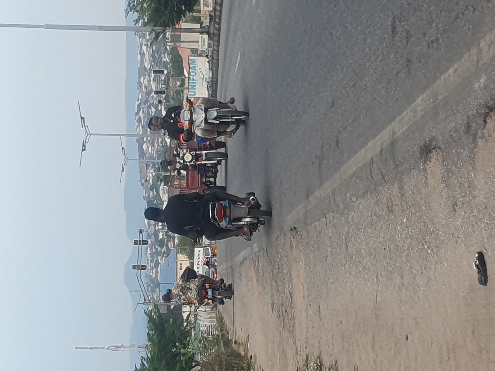 The image shows a street scene in Lokoja, Nigeria, with multiple motorcycles traveling on a paved road. Three motorcycles are prominently visible, each carrying a single rider. The riders are dressed in casual clothing. The road has a white line marking. Buildings and trees are visible in the background, suggesting an urban or suburban setting. In the far background, there is a large sign that says "UNIFOAM". There are also traffic lights and street lamps visible. The image appears to be taken during the daytime, with clear skies.