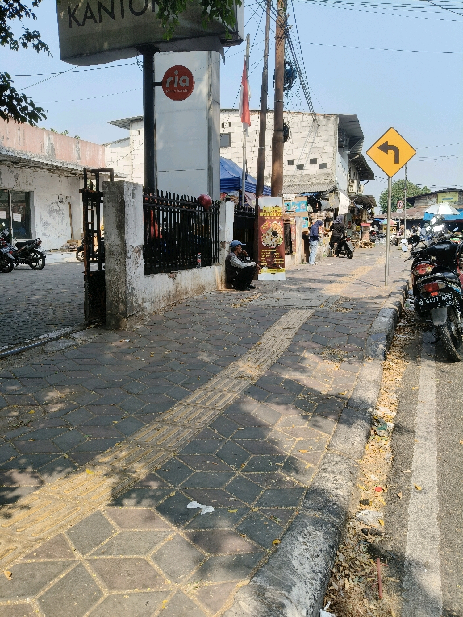 In the city of South Tangerang, Indonesia, on a bright, sunny day, a street scene unfolds. The foreground features a paved sidewalk with a textured pattern of diamond-shaped tiles and raised tactile paving indicators for the visually impaired. Scattered leaves litter the sidewalk and a concrete curb. To the right, a paved road with a white line marking is visible, with several motorcycles parked along its edge, one prominently displaying the license plate "B 6497 NEE".
In the middle ground, a man wearing a blue cap and dark clothing sits on the ground leaning against a concrete pillar. He is positioned near a black metal fence that encloses a building. Behind the fence, there are more motorcycles. A sign with a red circular logo and the text "ria" and "Money Transfer" is attached to a pole. Further back, a street market or small shops line the road, with vendors and customers visible. A yellow diamond-shaped traffic sign with a black arrow indicating a left turn is mounted on a pole. Buildings with white brick walls and corrugated roofs are visible in the background. An Indonesian flag hangs from a pole near a utility pole with numerous wires. The overall scene depicts a bustling urban environment with elements of commerce and daily life.