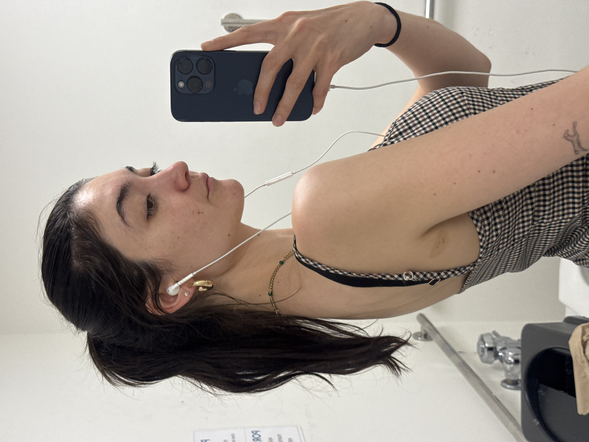 The image shows a young woman taking a mirror selfie in a public restroom. She is wearing a plaid dress and has her hair pulled back in a ponytail. She is also wearing white earbuds and gold hoop earrings. The restroom appears to be in a public building, possibly a community center or a government building. The location is New York City, United States of America.