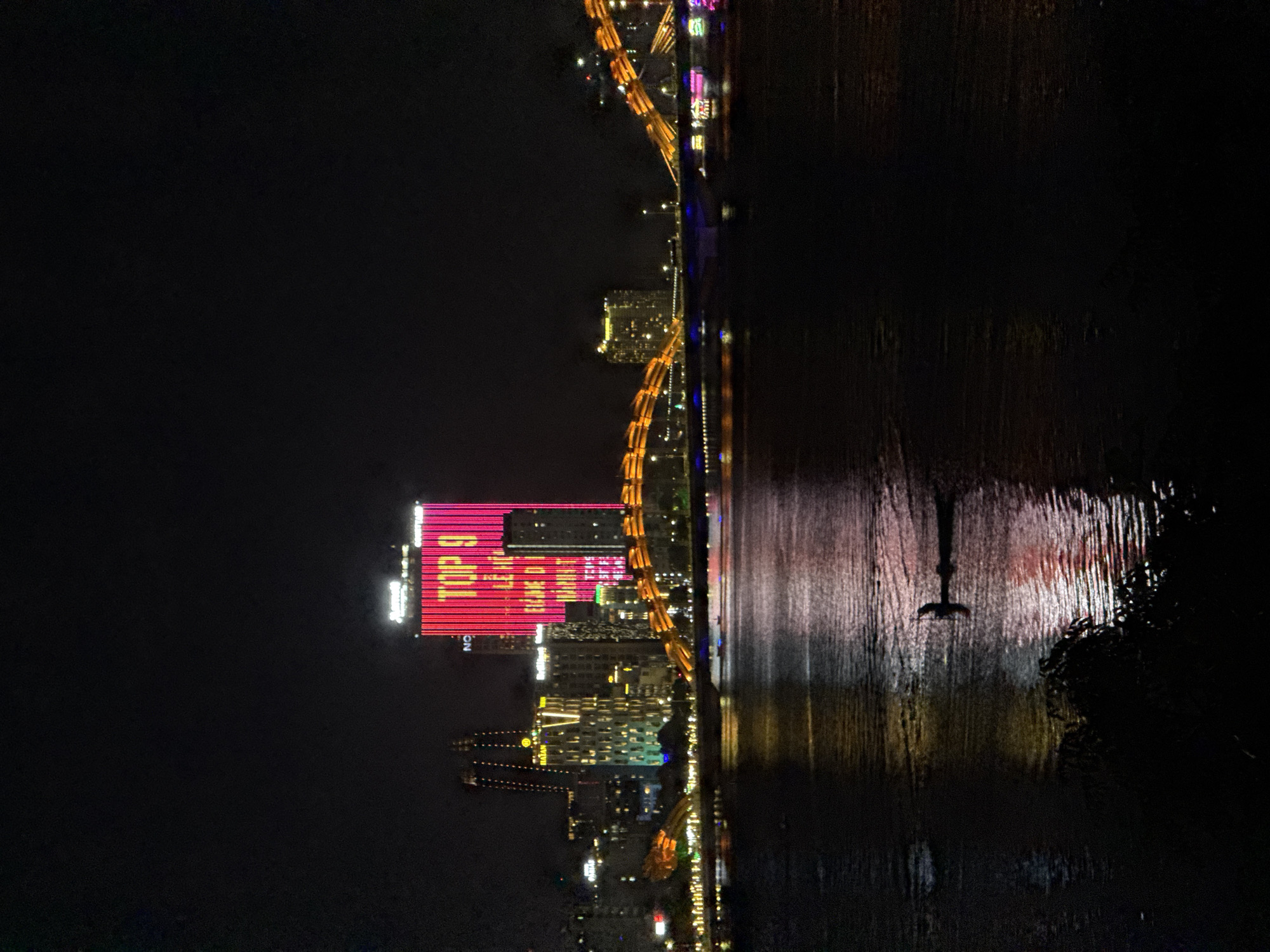 The image captures a nighttime cityscape of Da Nang, Vietnam, dominated by the illuminated Dragon Bridge and surrounding buildings. The foreground features a dark body of water, likely the Han River, reflecting the vibrant lights from the city. In the water, a small silhouette of a person in a boat is visible. The Dragon Bridge, a prominent landmark, is adorned with orange lights, arching across the river. Beyond the bridge, various buildings are lit up, including a tall skyscraper displaying a large pink and red advertisement with the text "TOP 9" and other illegible lettering. The overall scene is one of urban nocturnal activity, with the artificial lights creating a dramatic contrast against the dark night sky.